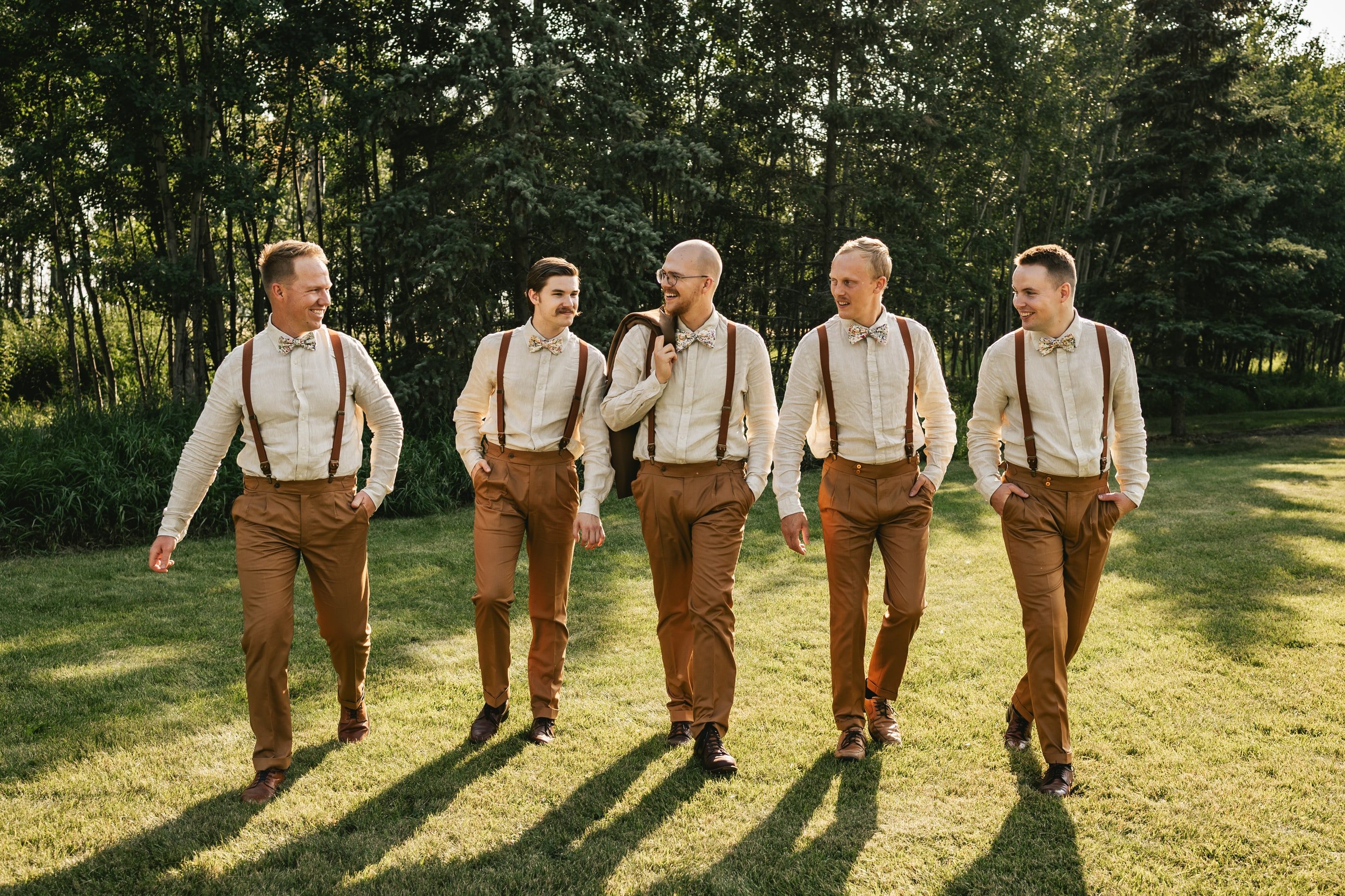 A groom and his groomsmen in white shirts, suspenders and brown trousers walk across a sunny lawn.