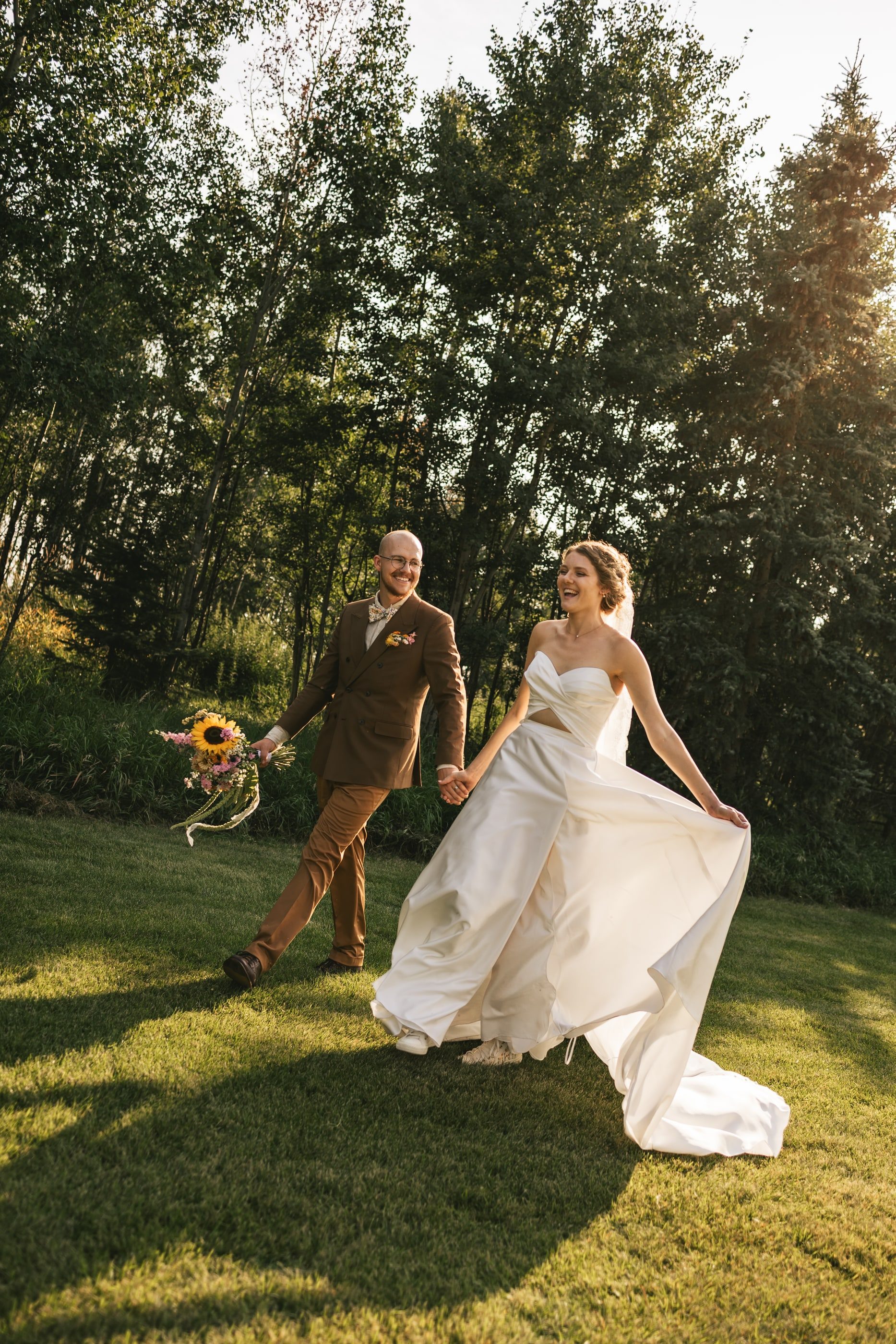 A bride and groom walk hand in hand across the lawn in evening sun, her train sweeping behind her.
