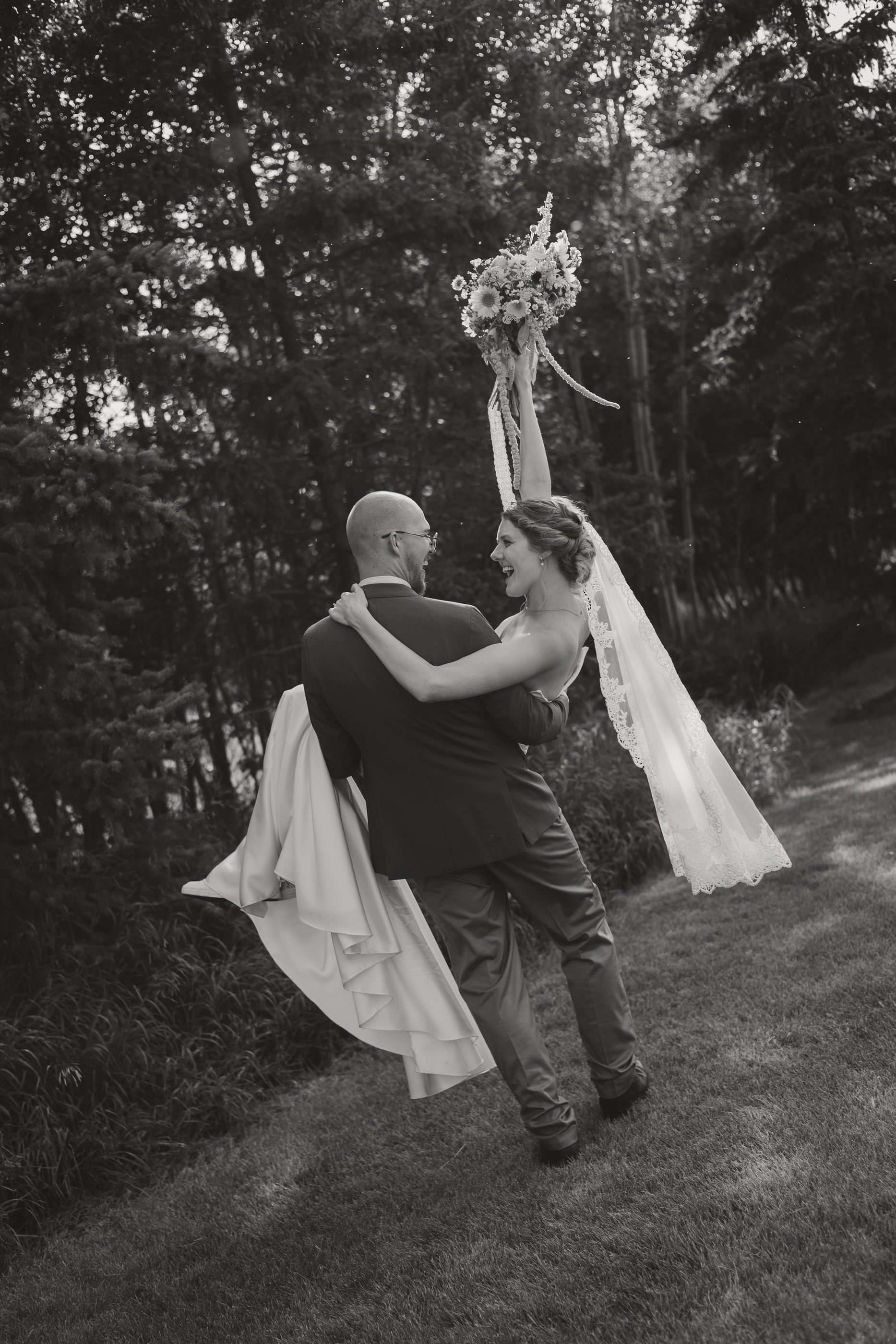 Black-and-white photo of a groom carrying his bride as she raises her bouquet in the air.
