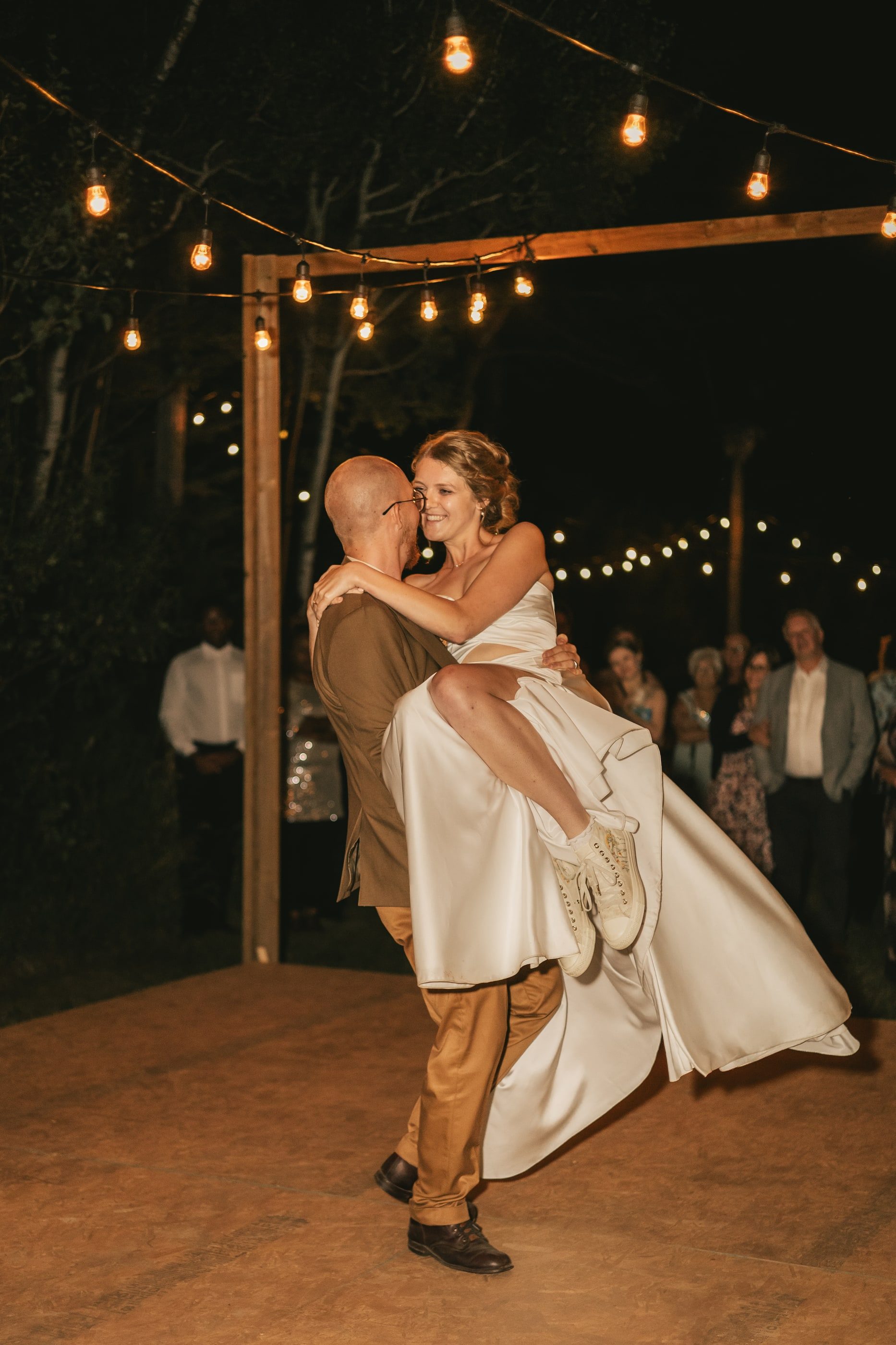 A groom lifts his bride on a dance floor beneath strings of lights.