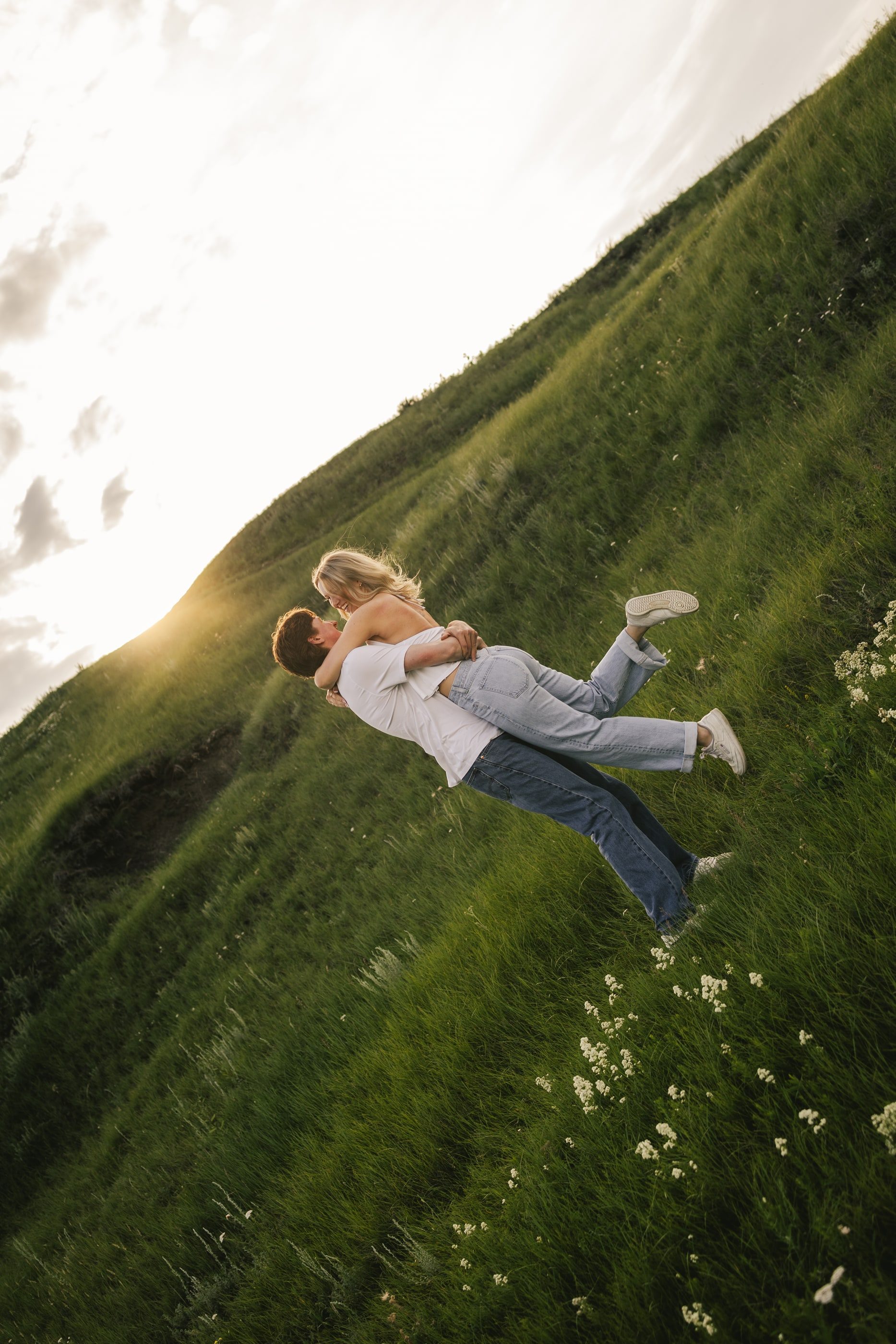 A man lifts his laughing partner on a steep green hillside, the sun low behind them.