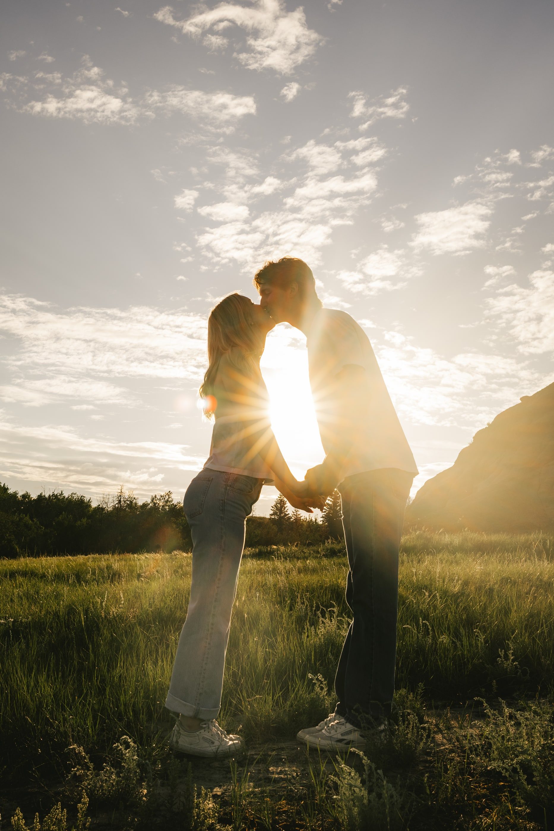 A couple leans in for a kiss, silhouetted against the sun in a grassy valley.