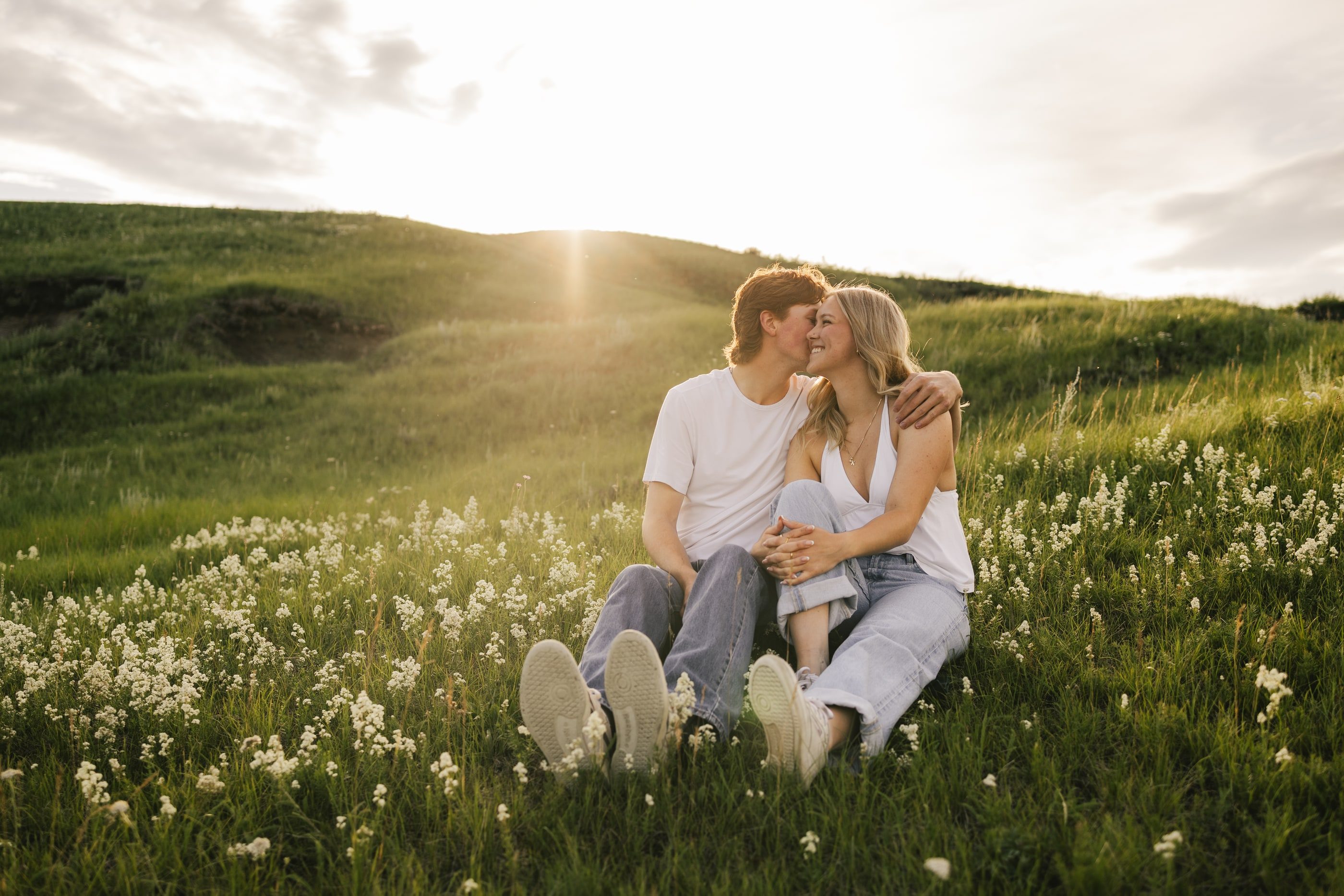 A couple sits close and kisses among white wildflowers on a sunlit hillside.