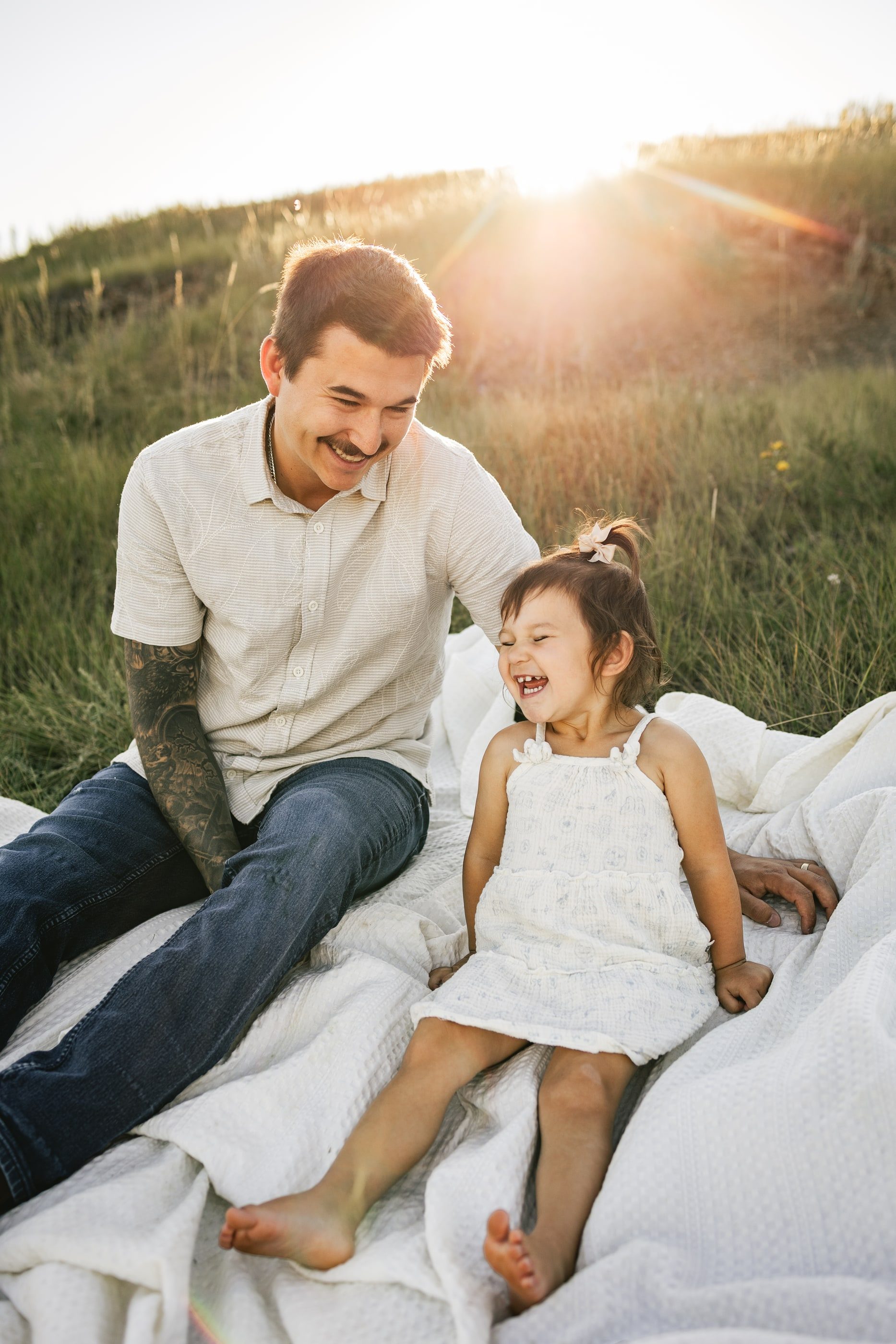 A father laughs with his toddler daughter on a white blanket in a sunlit grassy field.