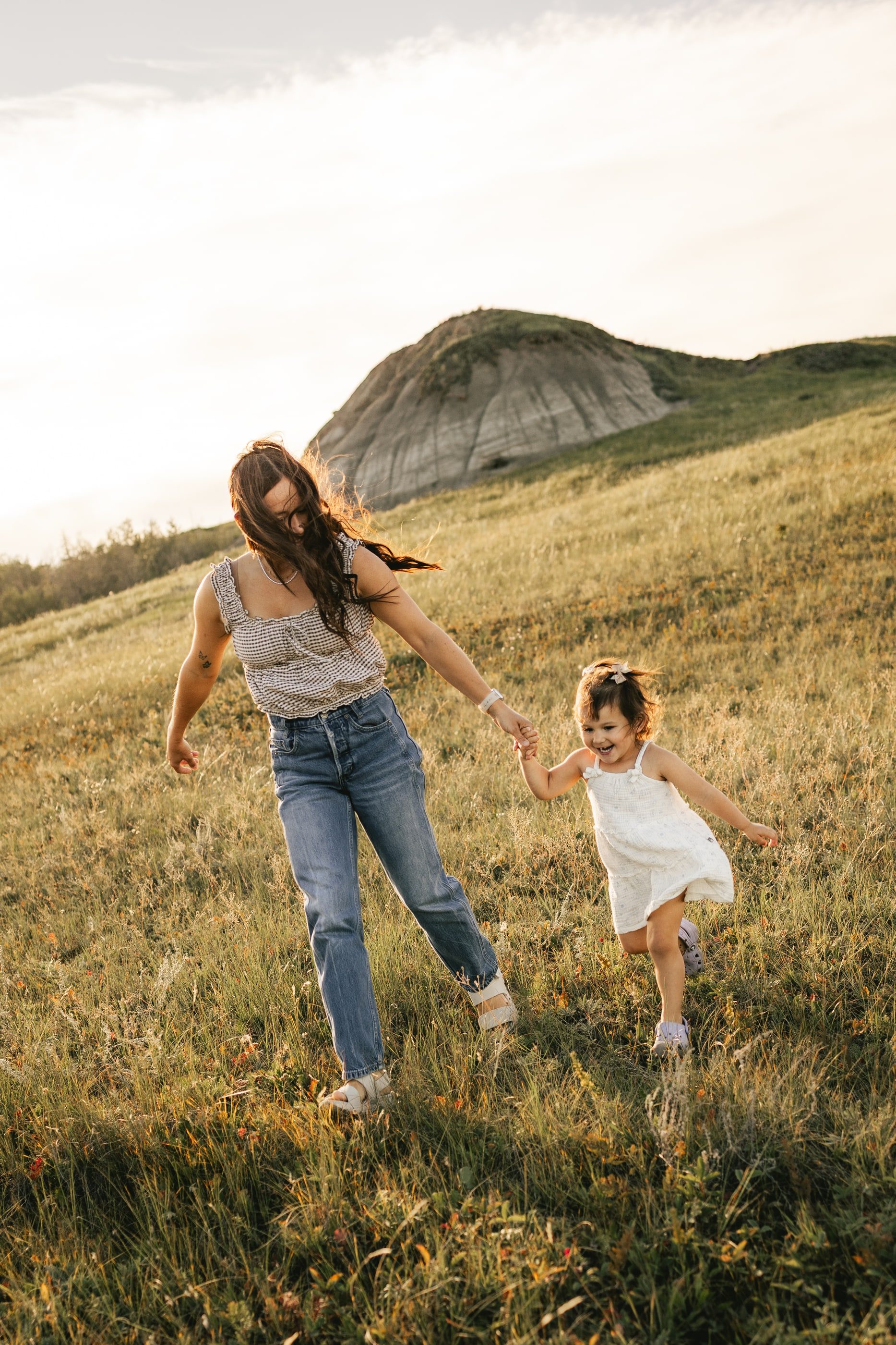 A mother leads her little girl by the hand across a grassy hillside below a rounded badlands butte.