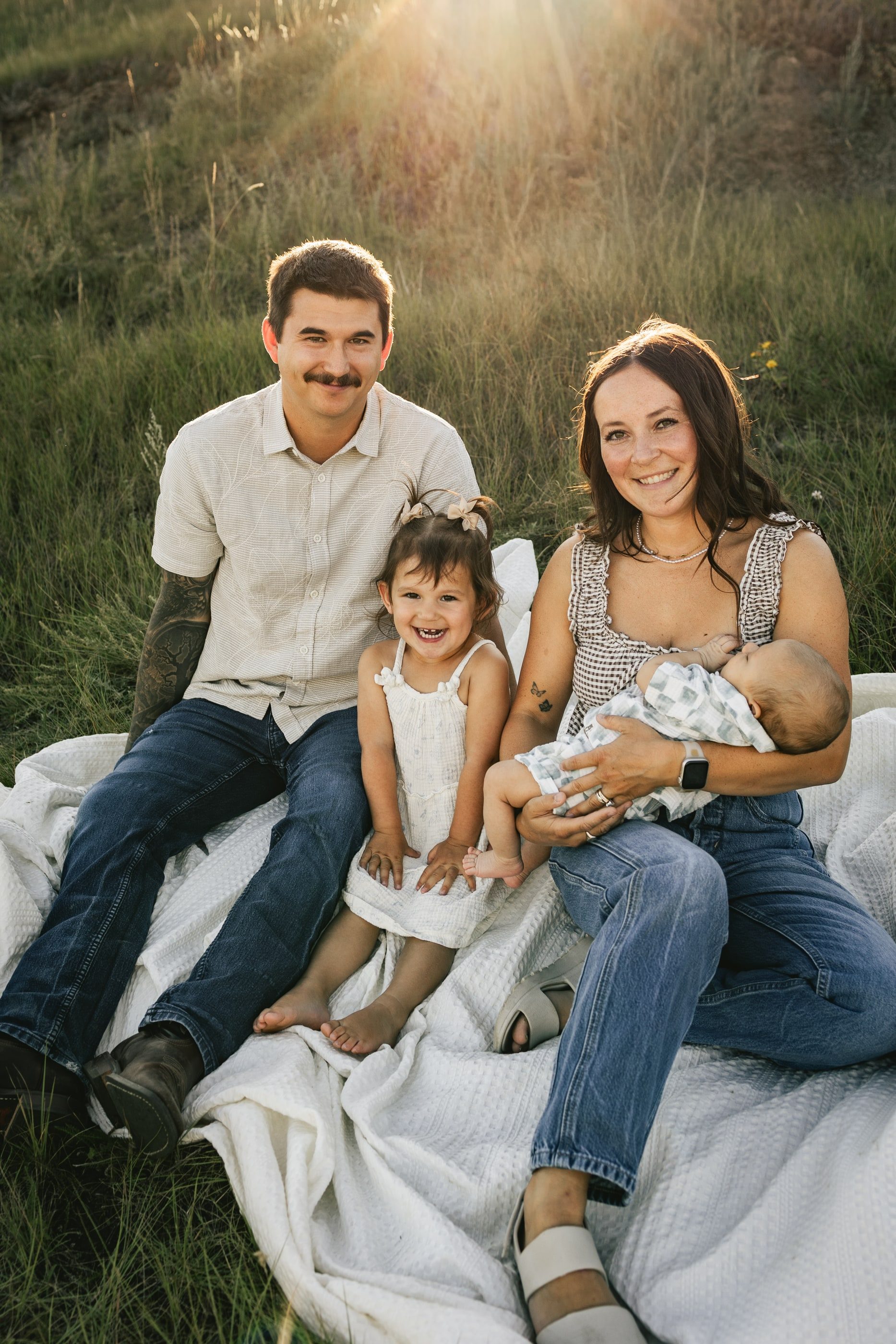 Parents sit on a blanket in the grass with their toddler and newborn, golden light behind them.