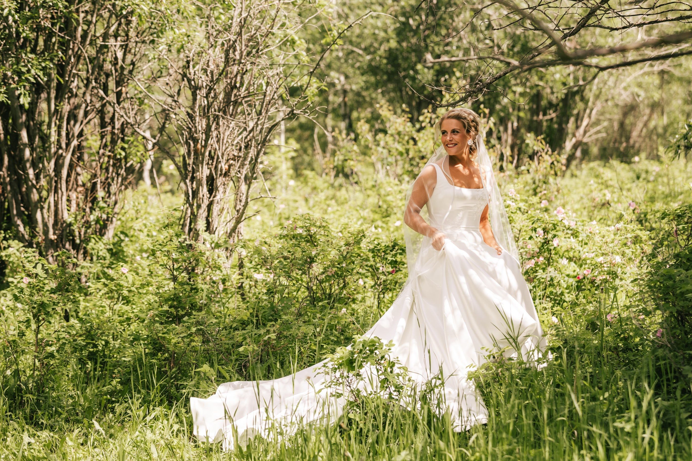 A bride walks through tall green grass in a wooded meadow, her gown trailing behind her.