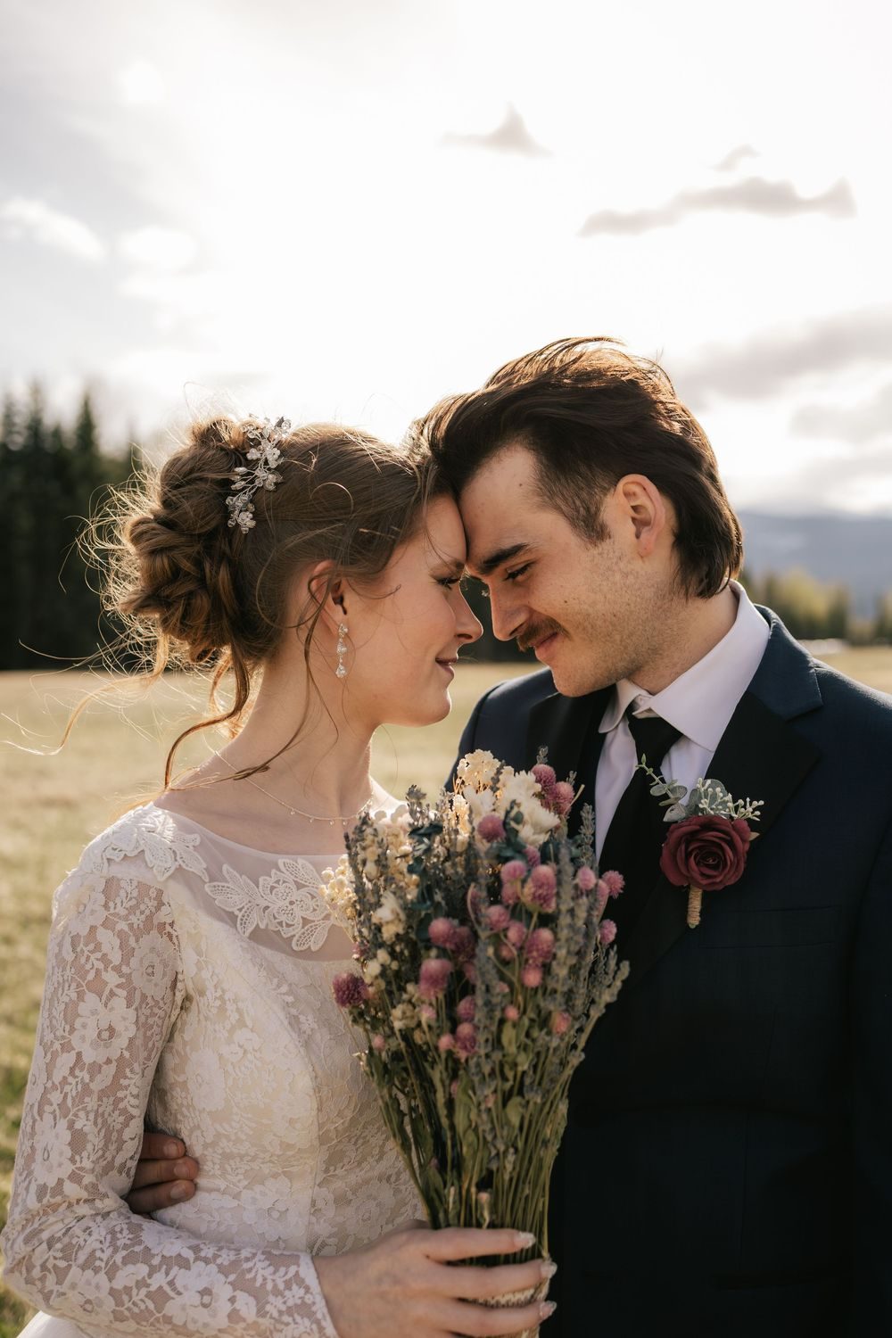 A bride and groom rest their foreheads together, her bouquet of dried wildflowers between them.