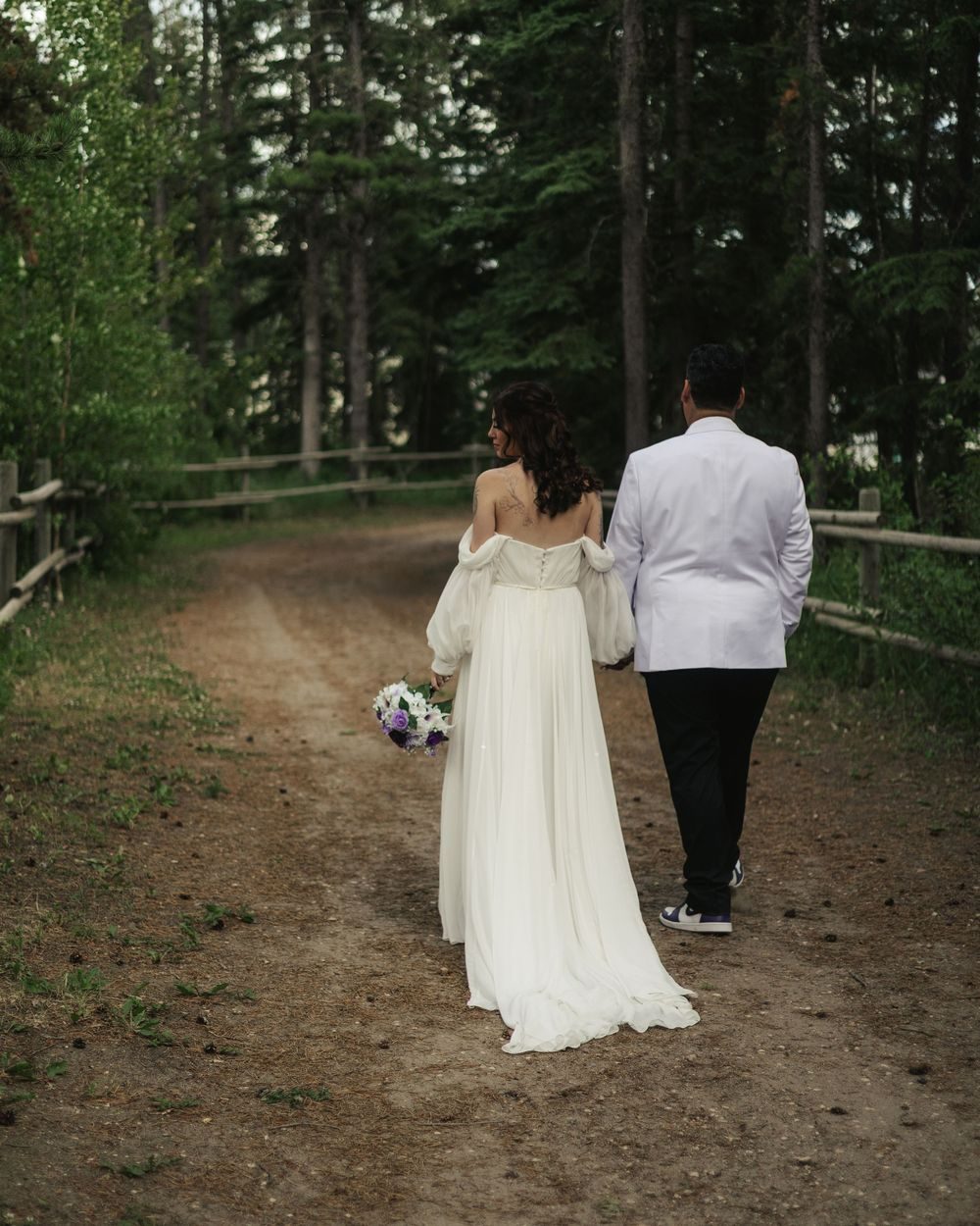 A bride and groom walk away together down a quiet forest path.