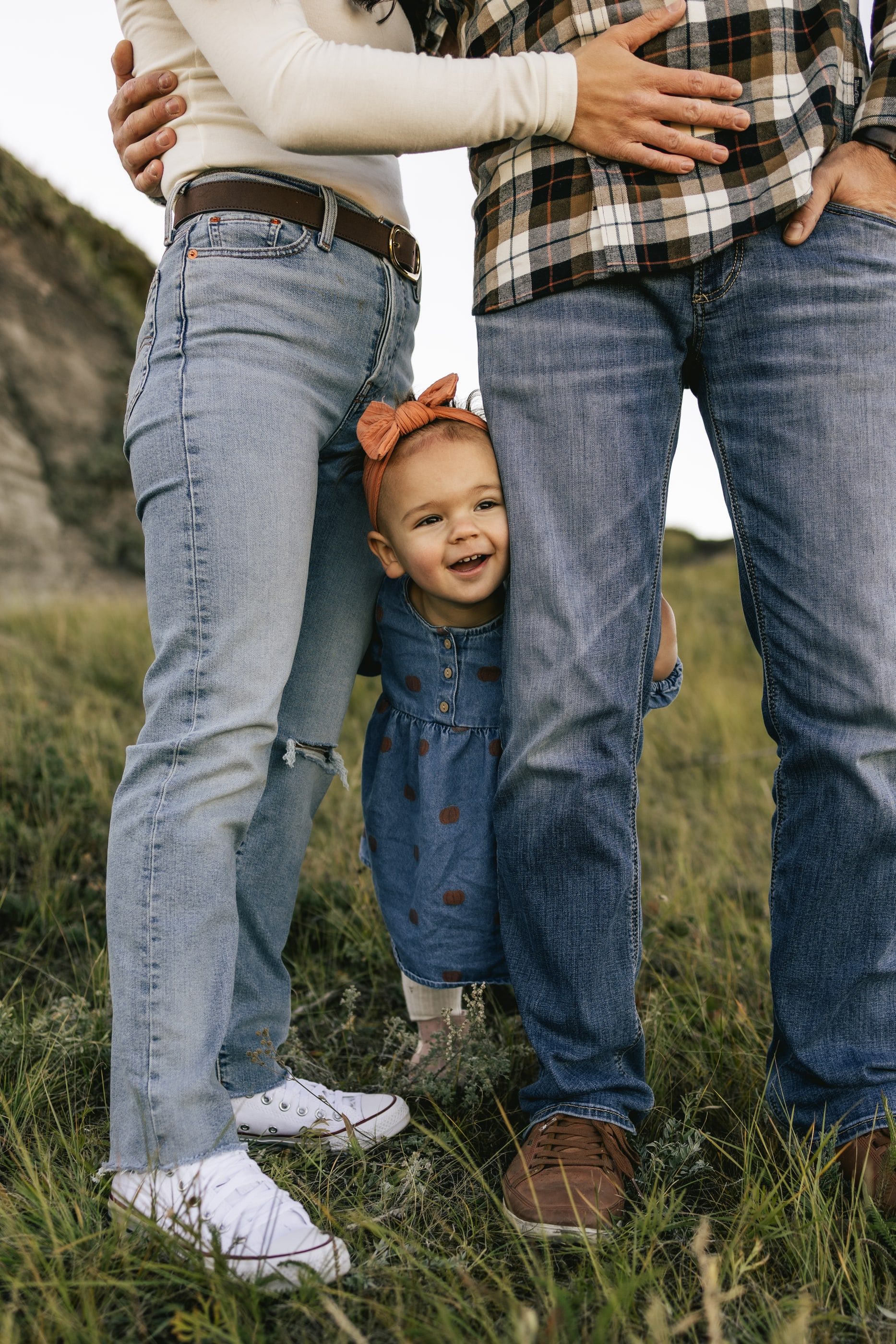 A laughing toddler with a bow in her hair peeks out between her parents' legs.