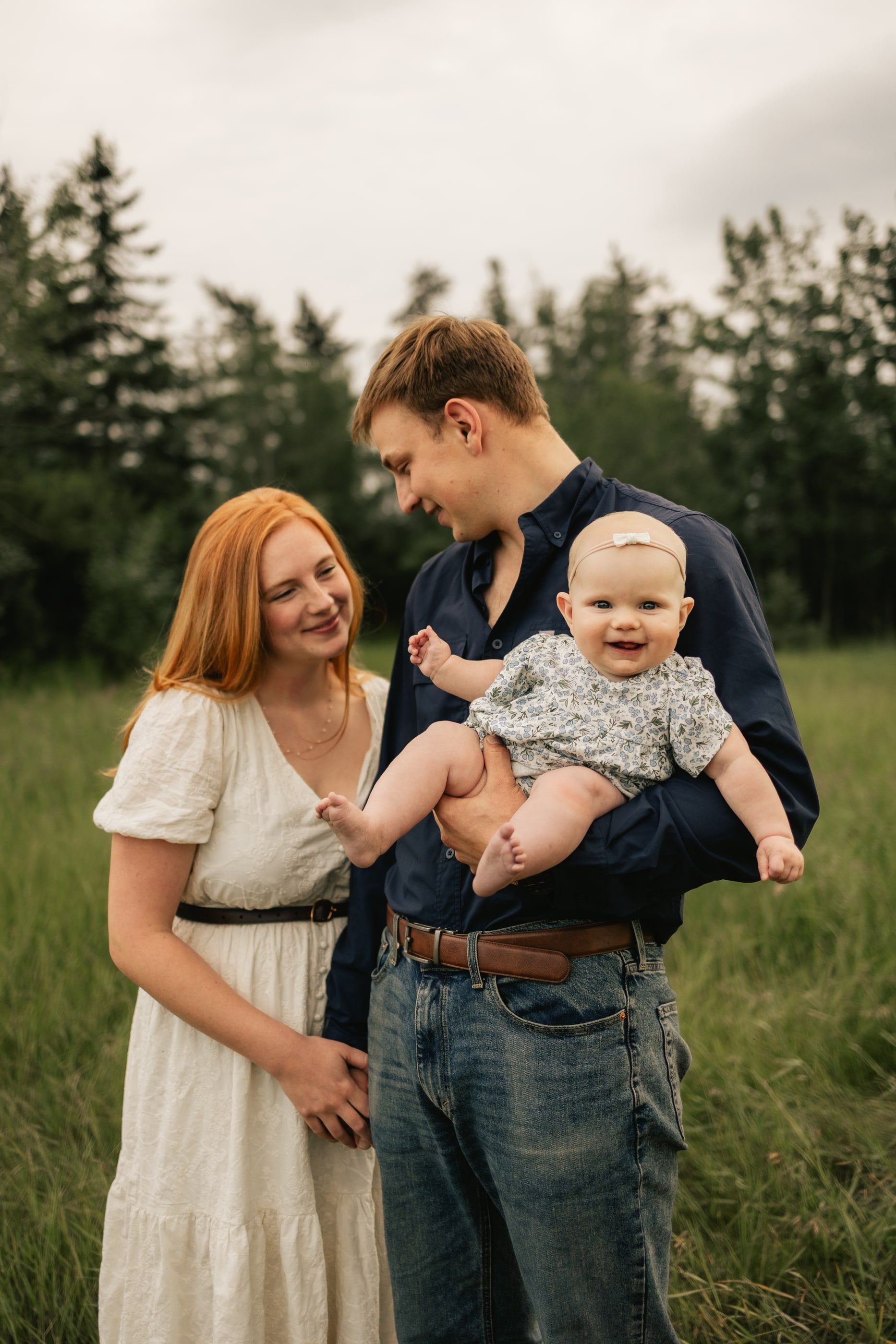 A father holds up their laughing baby as the mother smiles beside him in a grassy meadow.