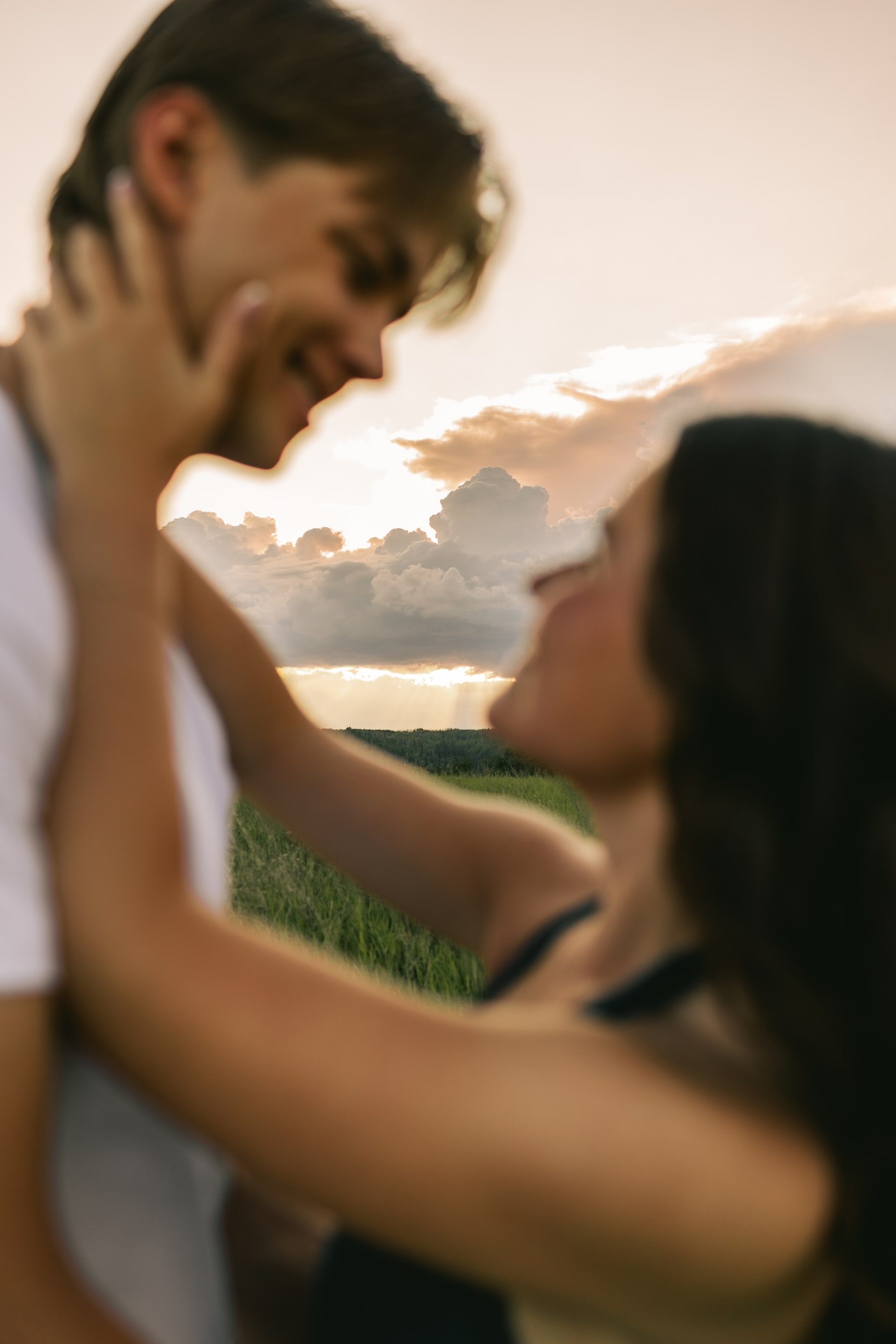 A couple laughs face to face, her hand on his cheek, against a glowing evening sky.