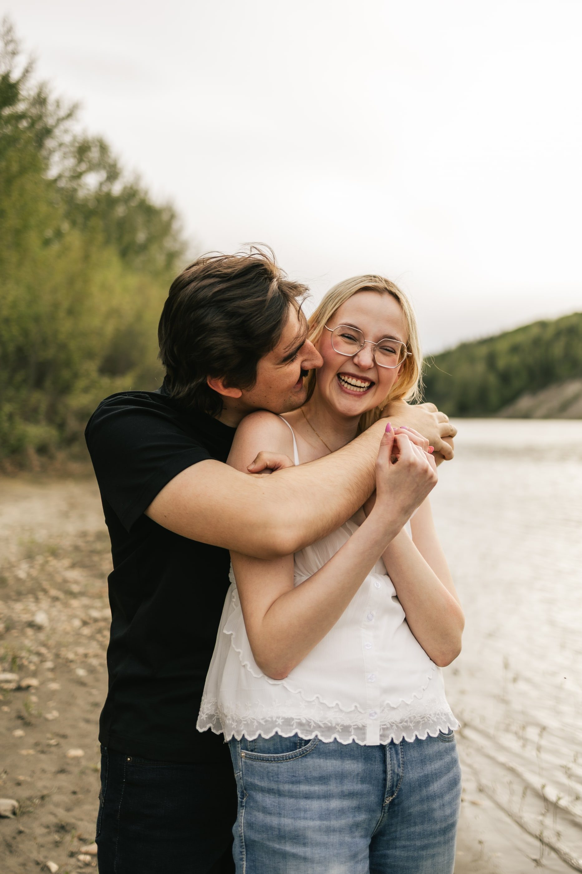A man hugs his laughing partner from behind and kisses her cheek on a riverbank.