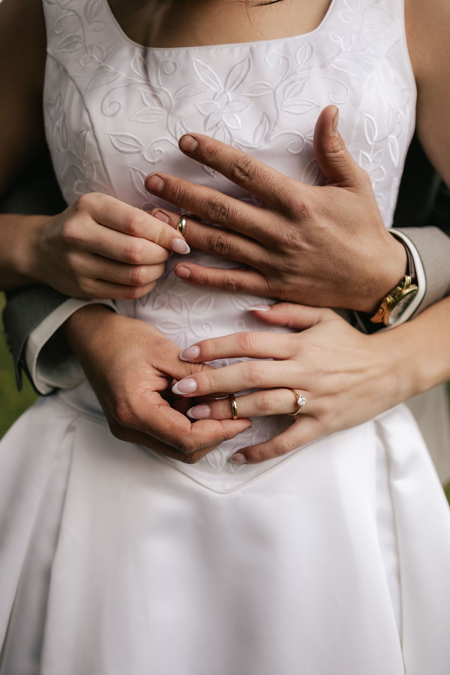 A groom's hands rest at the bride's waist, her hands over his, wedding rings catching the light.