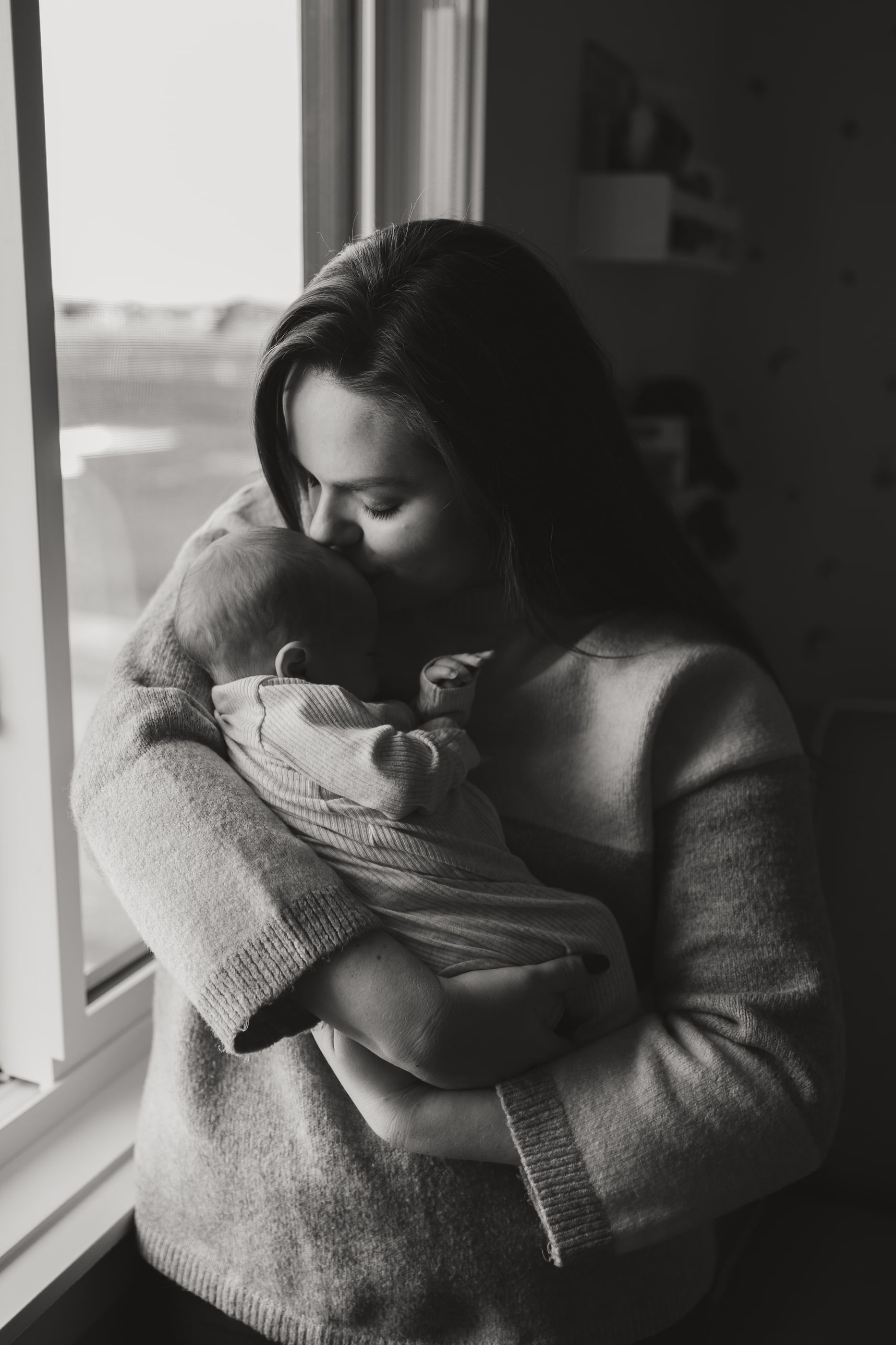 Black-and-white photo of a mother kissing her newborn by a window.