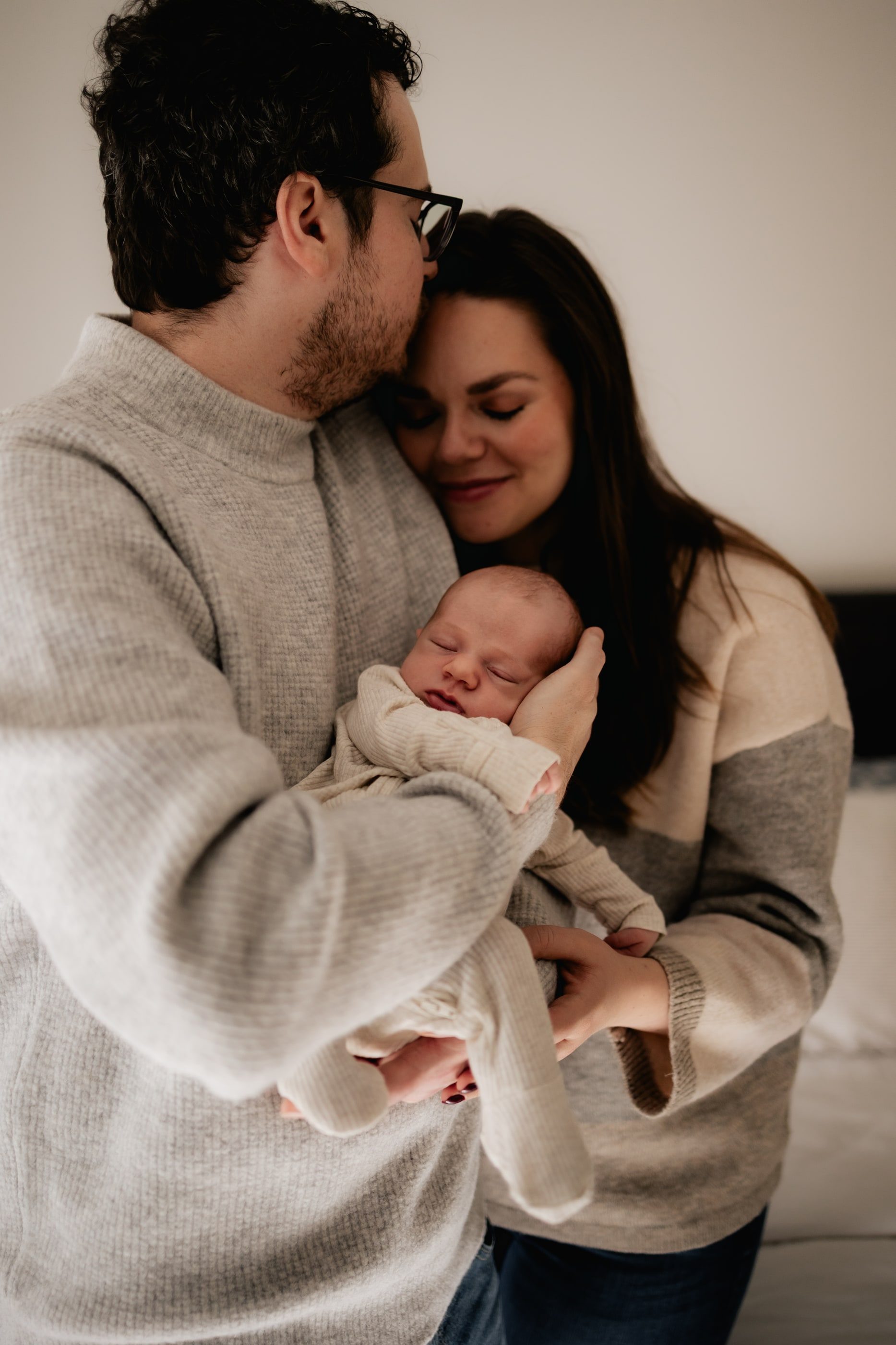 A father kisses the mother's forehead as she holds their sleeping newborn.