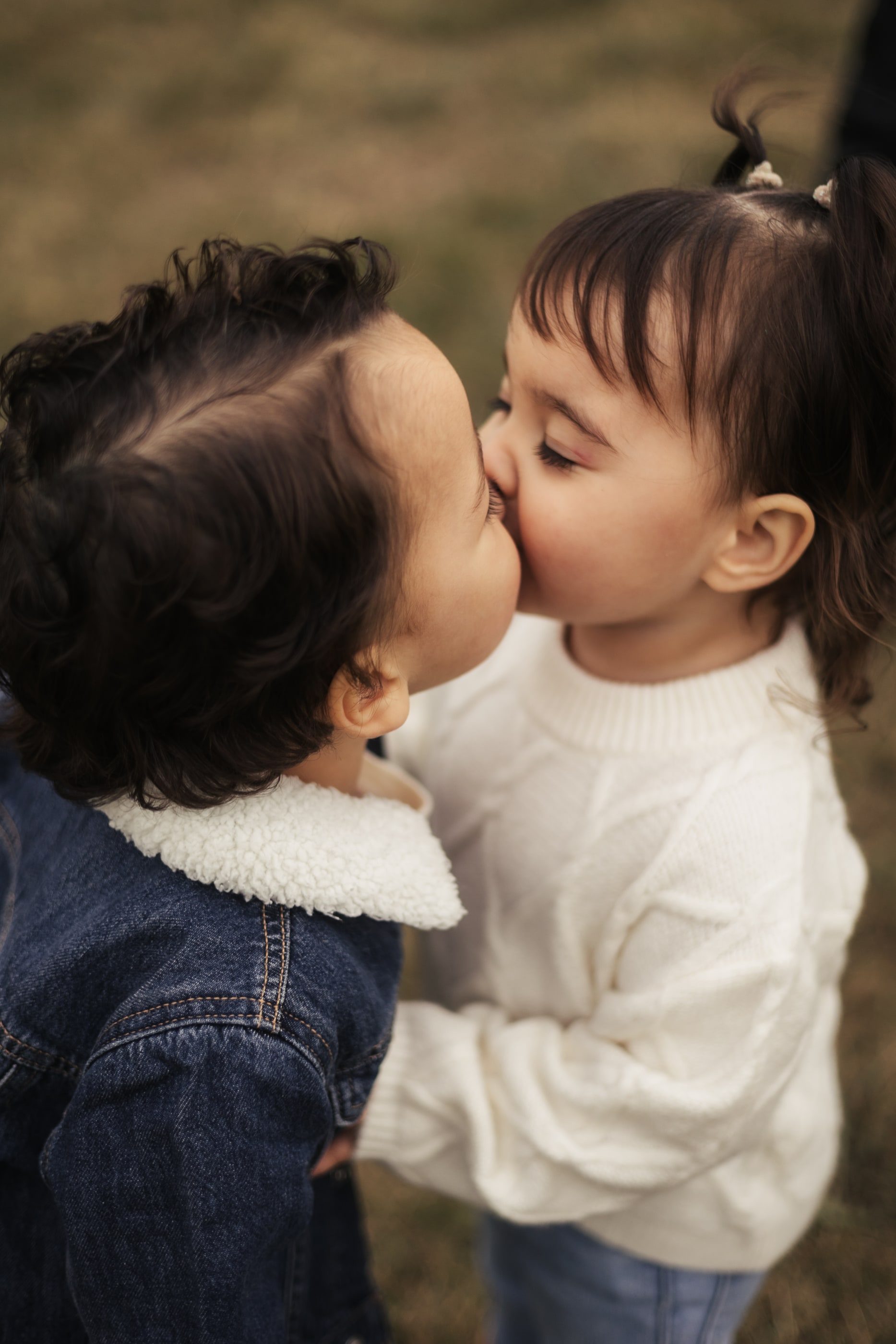 Two toddlers, one in a denim jacket and one in a white sweater, share a kiss.