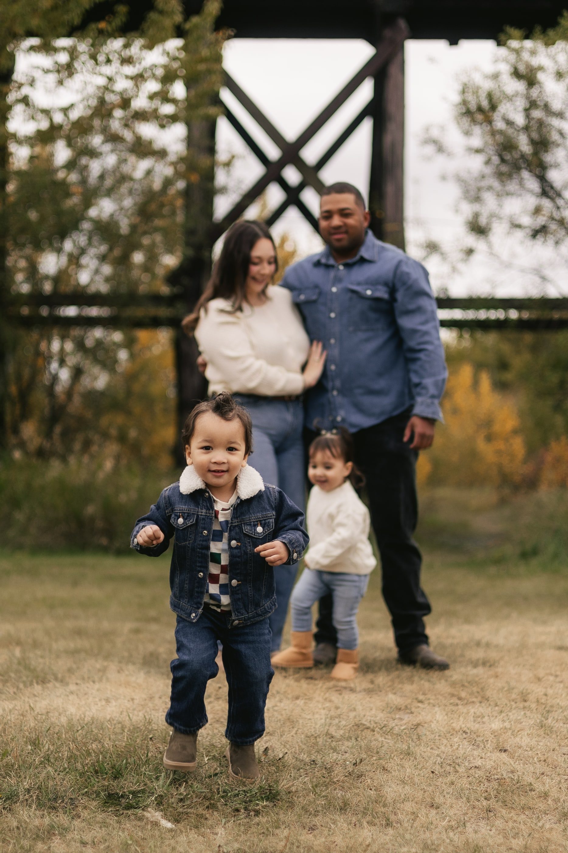 A toddler in a denim jacket runs toward the camera as his family stands behind him beneath a railway trestle.