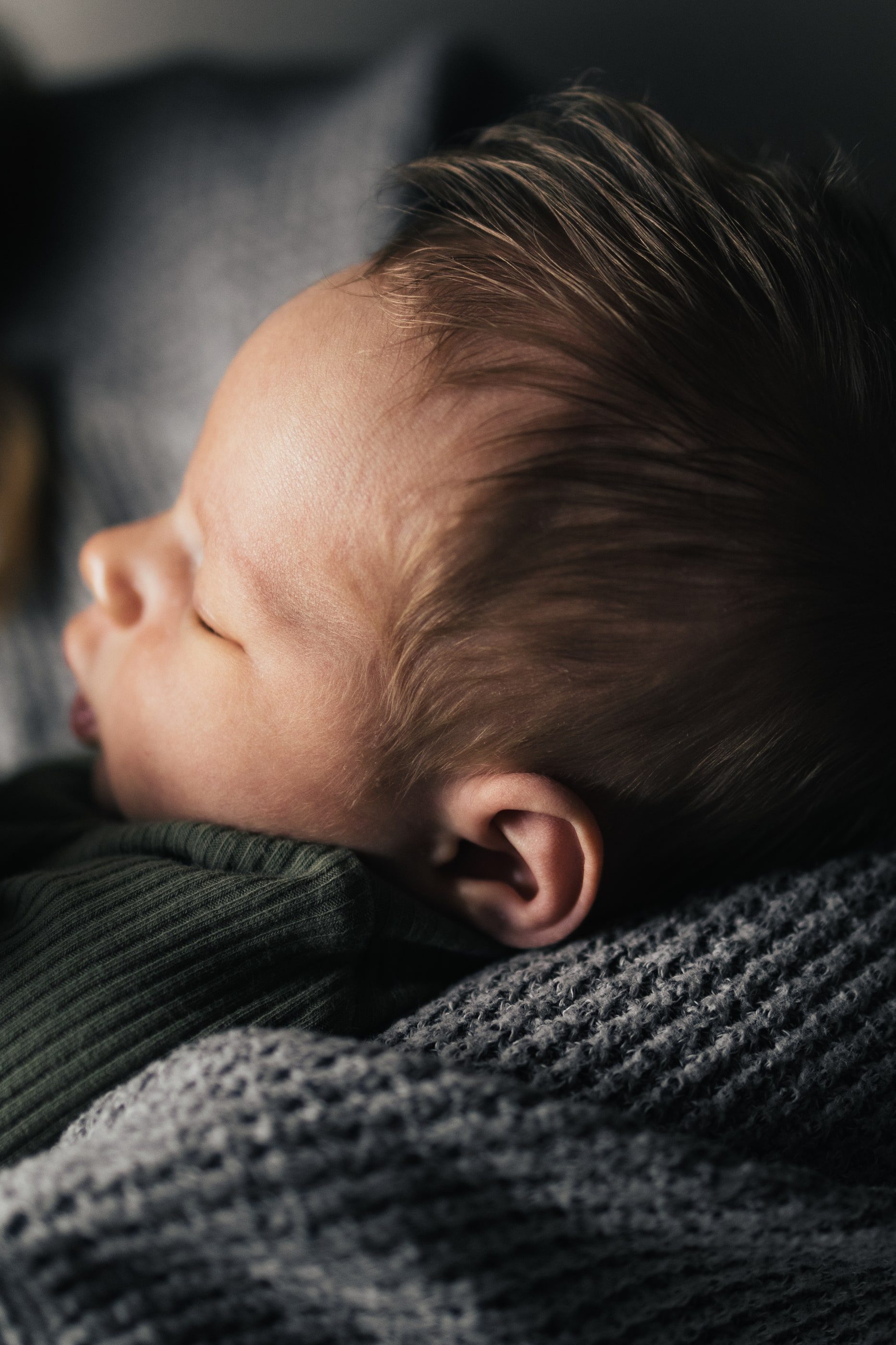 Close-up profile of a sleeping newborn's face and soft hair against a grey knit blanket.