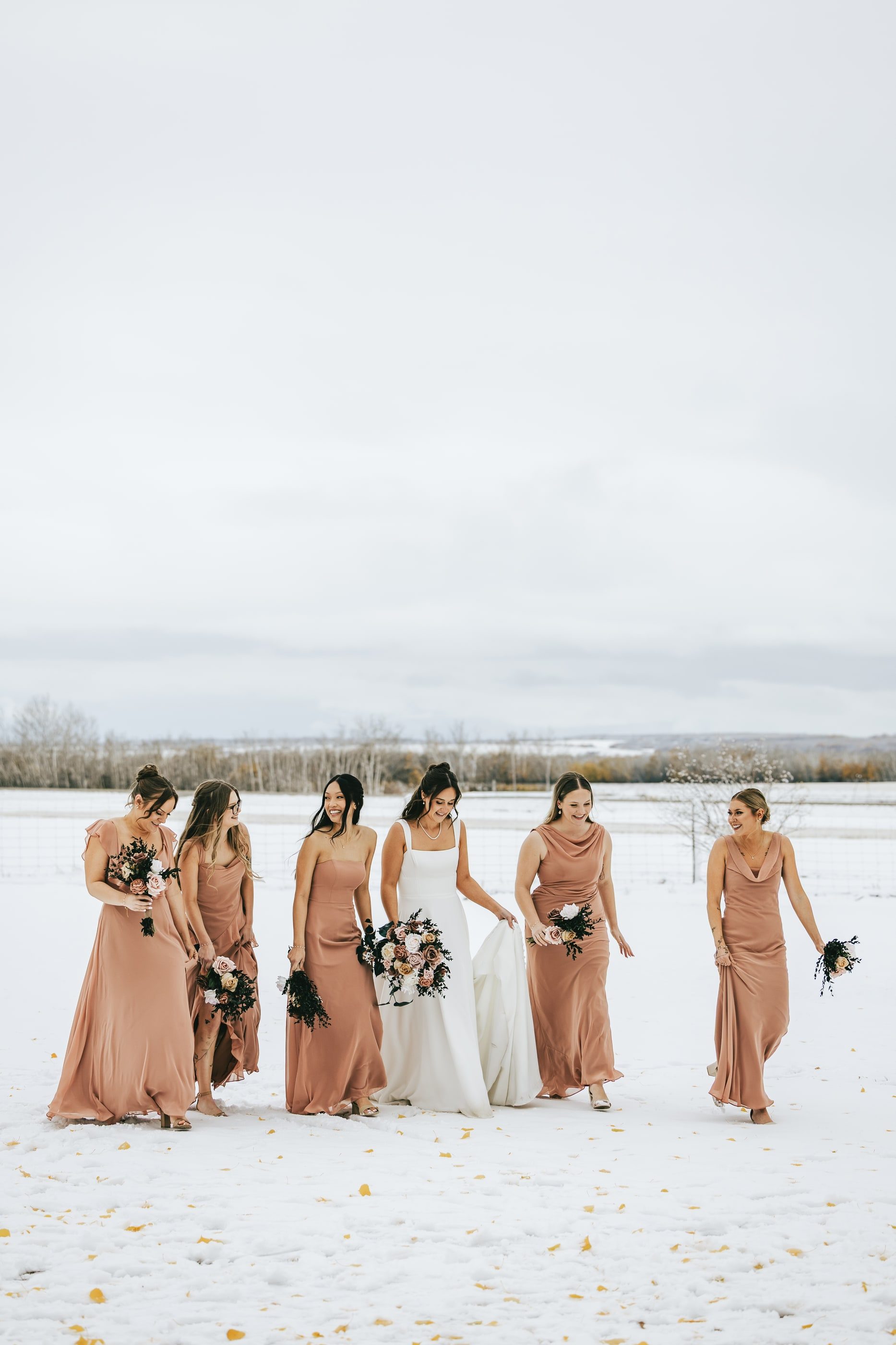 A bride and her bridesmaids in blush dresses walk across a snowy field.