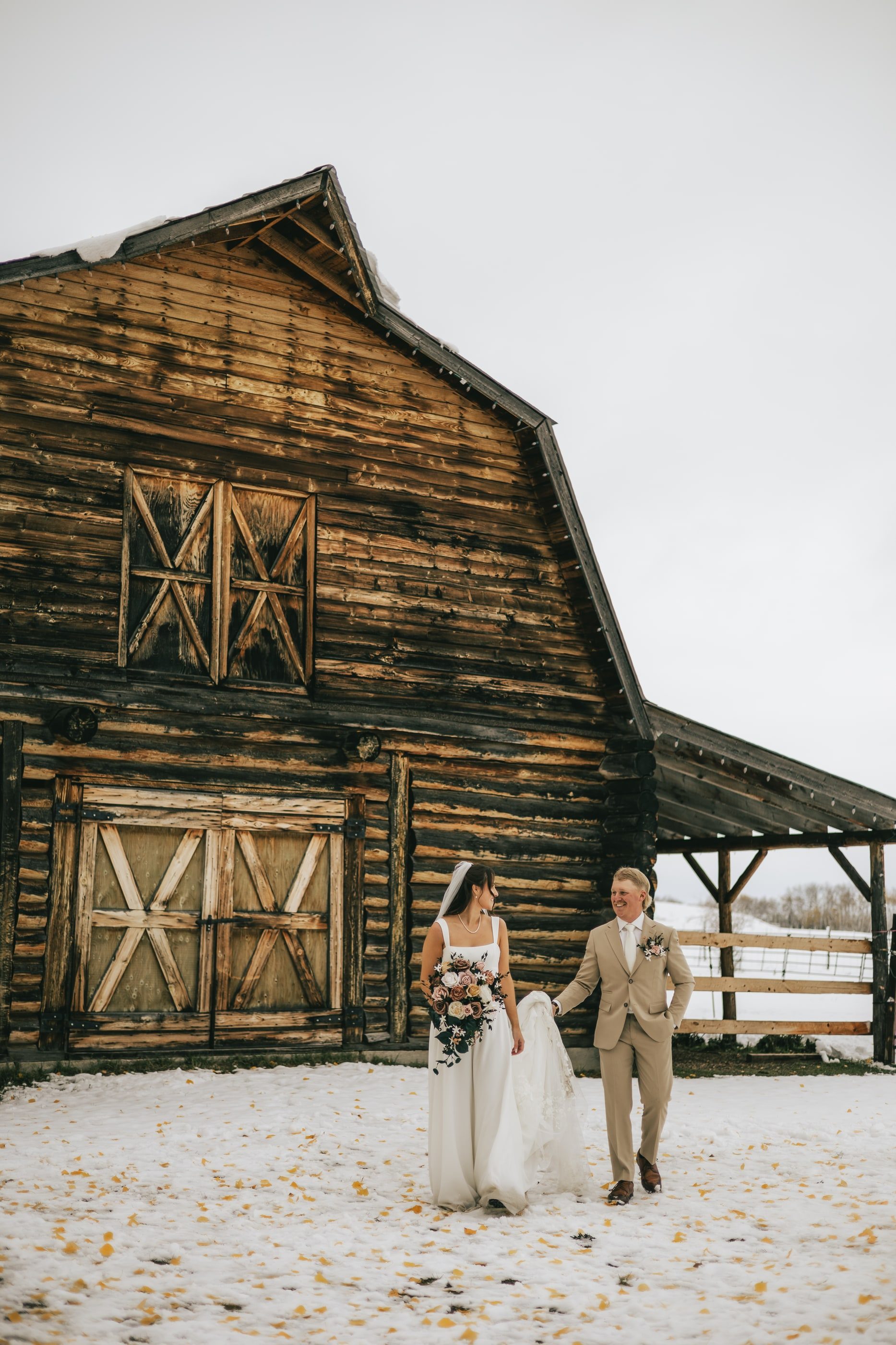 A bride and groom walk arm in arm past a weathered wooden barn on a snowy day.