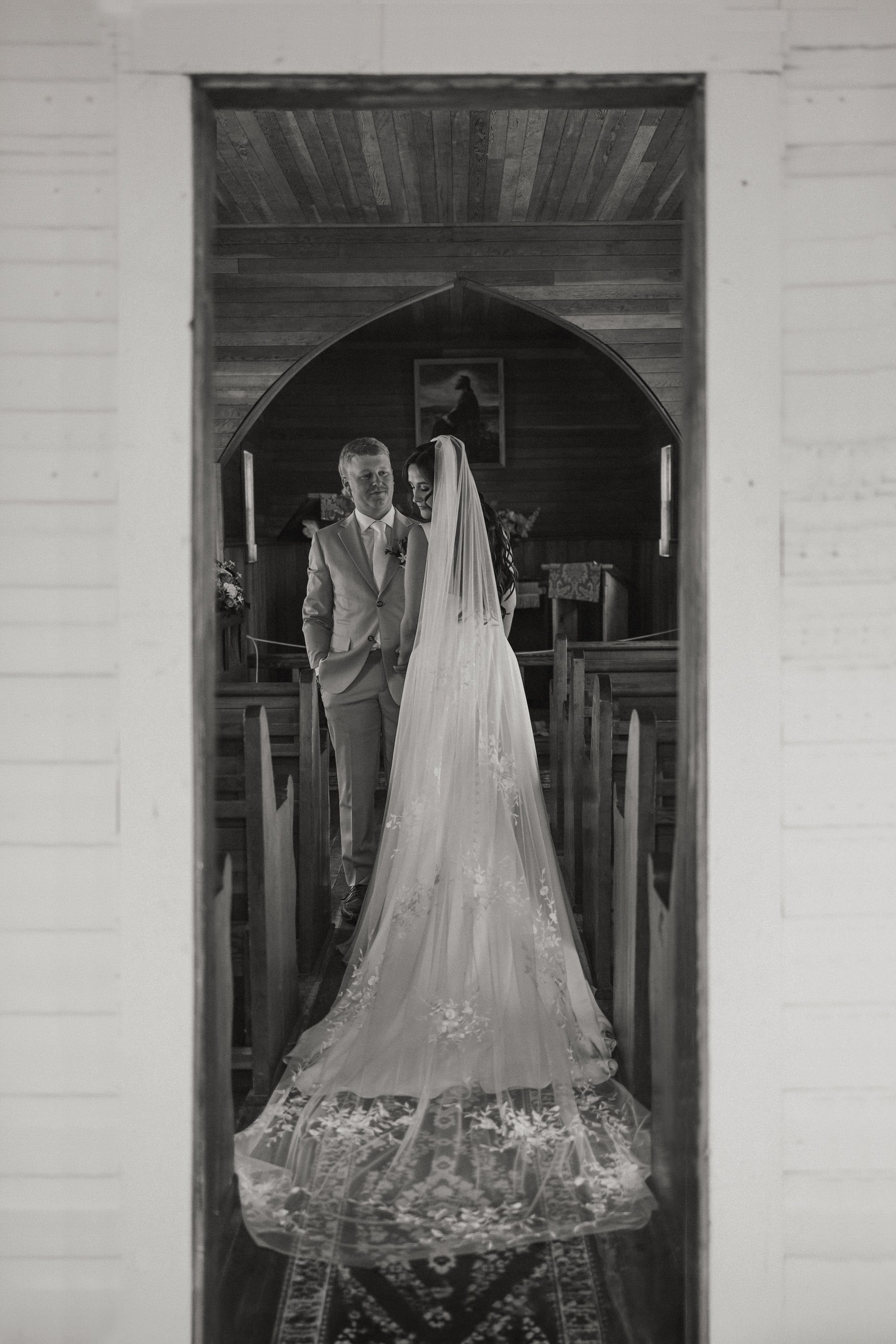 Black-and-white photo of a bride's long lace veil trailing behind her at the altar of a small chapel.