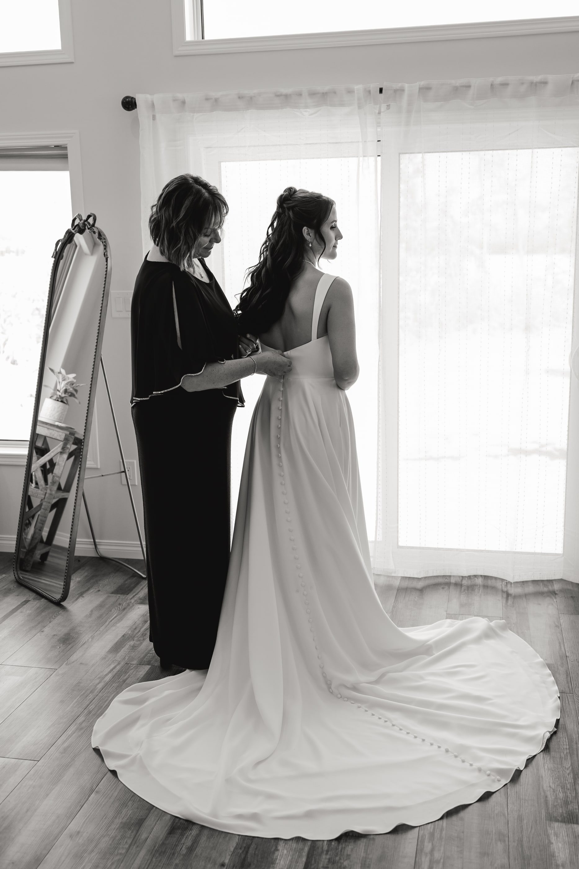 Black-and-white photo of a mother fastening the back of her daughter's wedding dress by a bright window.