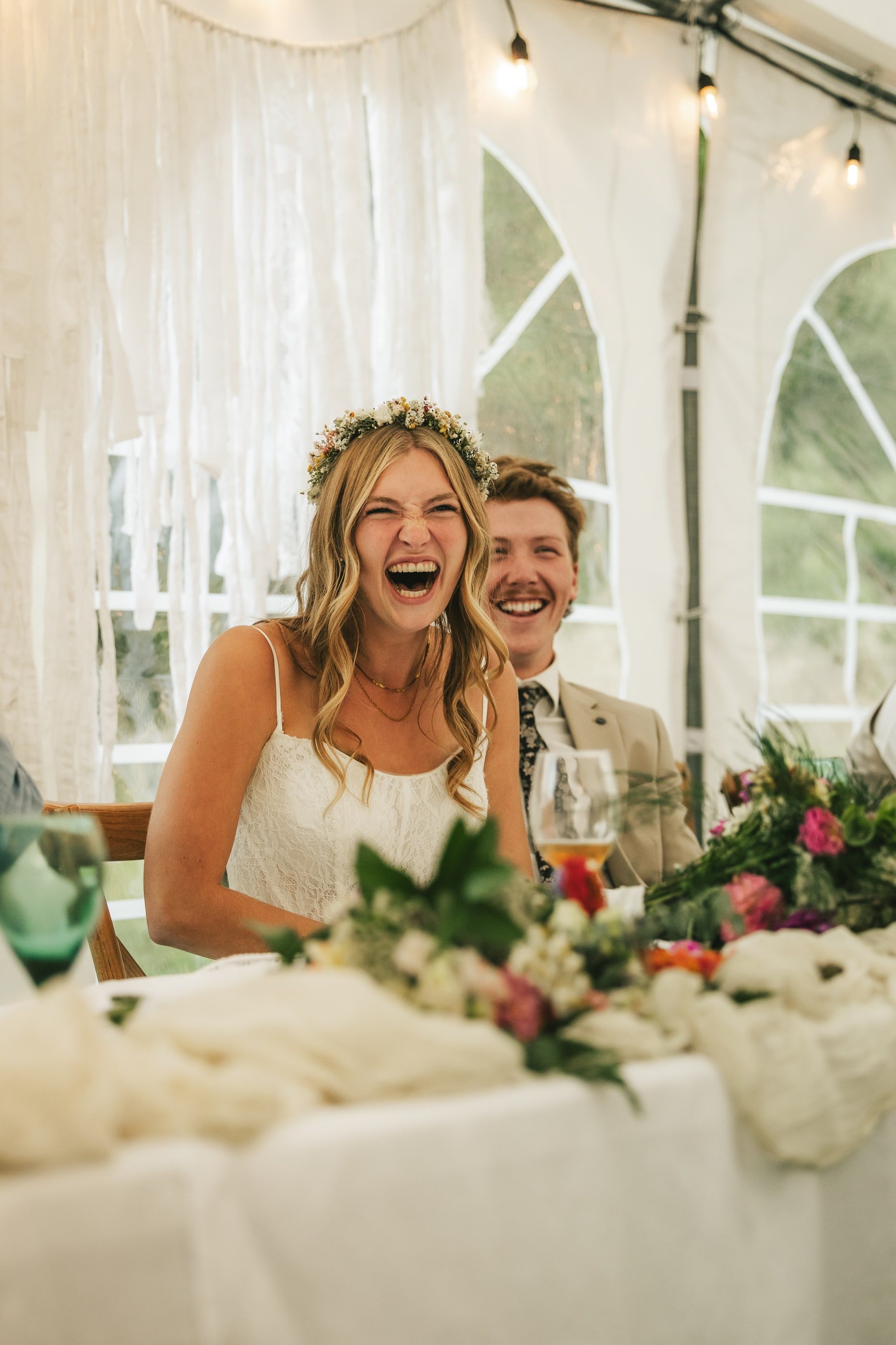 A bride in a flower crown laughs with her groom at the head table under a white tent.
