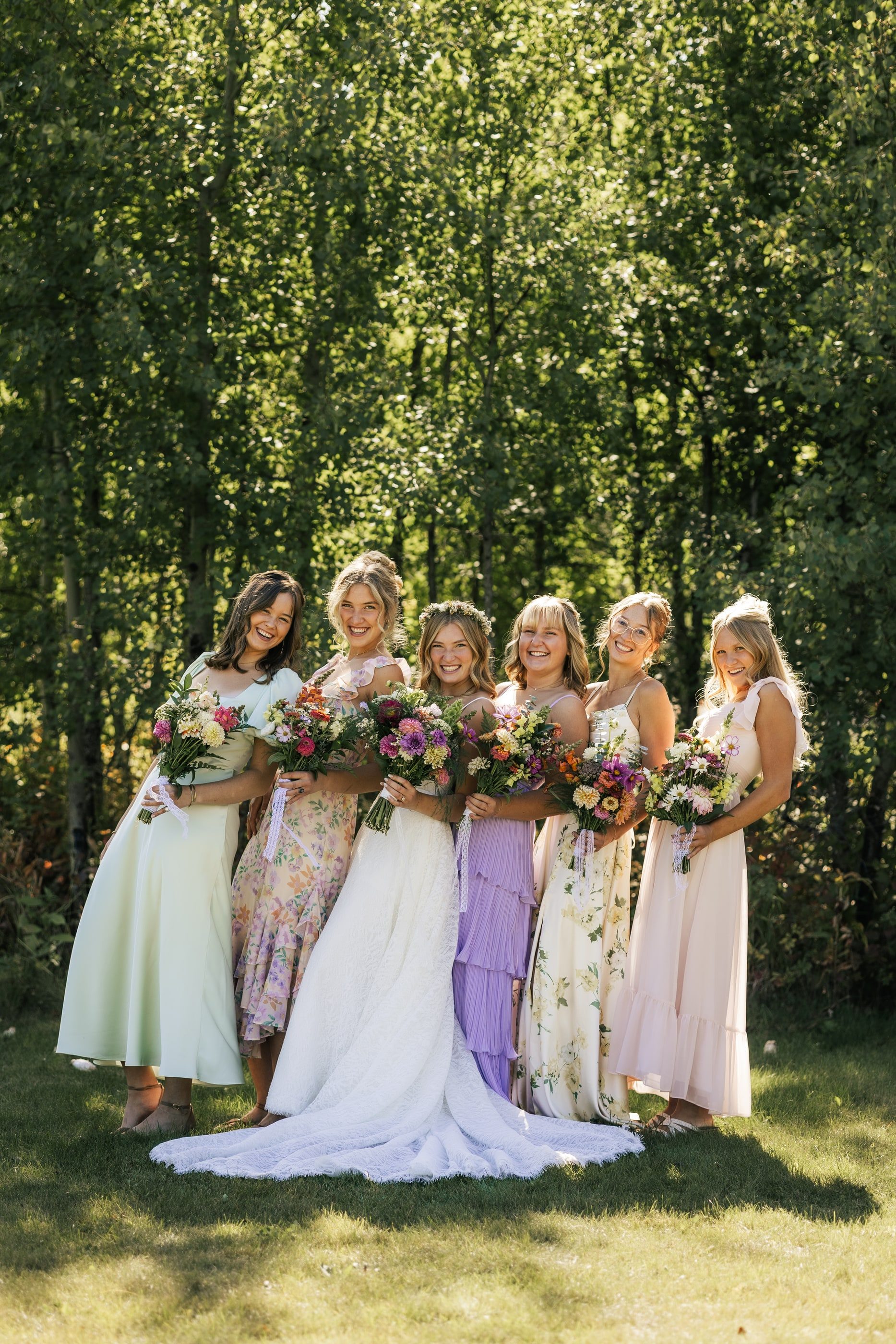 A bride stands with her bridesmaids in pastel and floral dresses, all holding colourful bouquets among the aspens.
