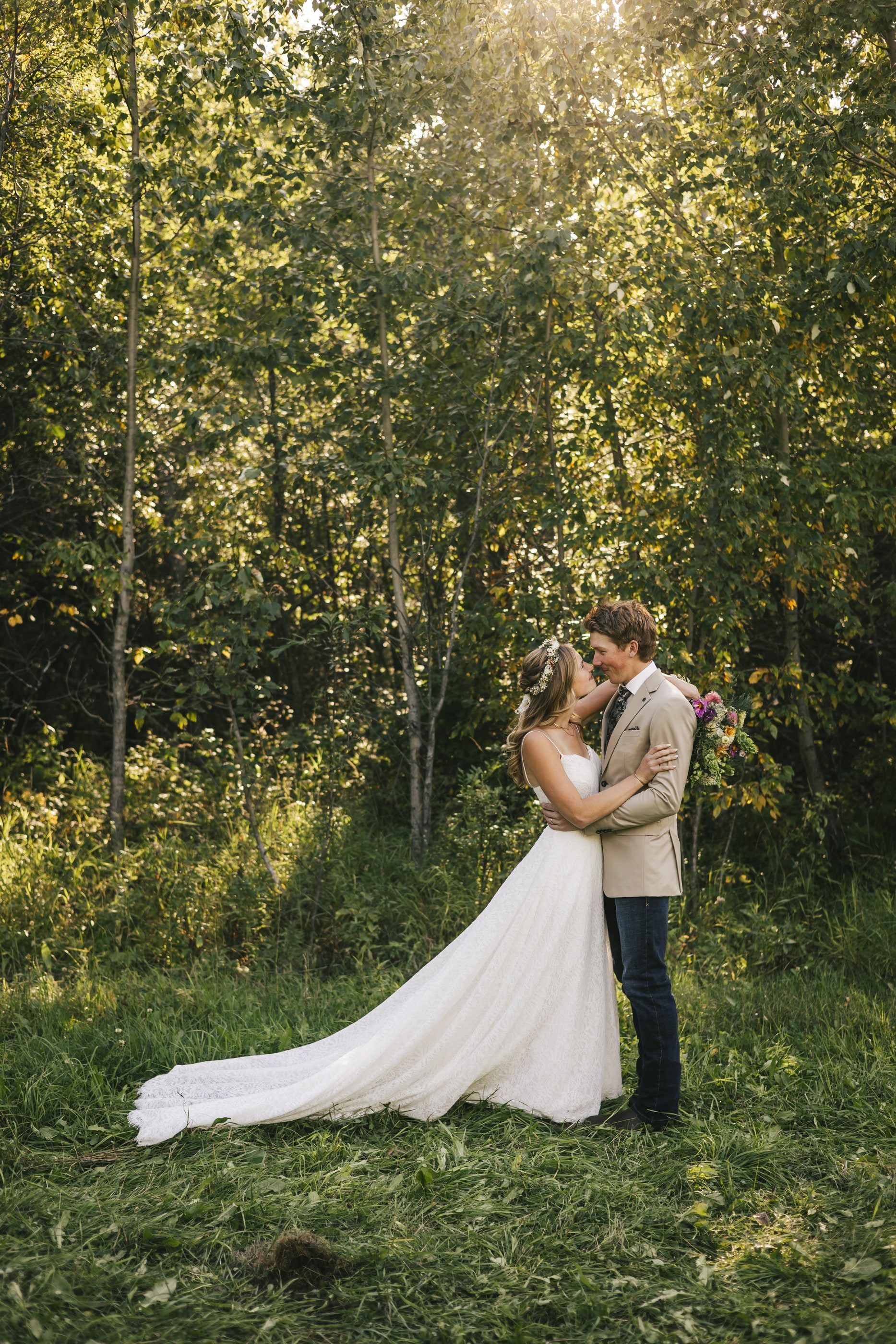 A bride and groom kiss in a sunlit aspen clearing, her train spread across the grass.