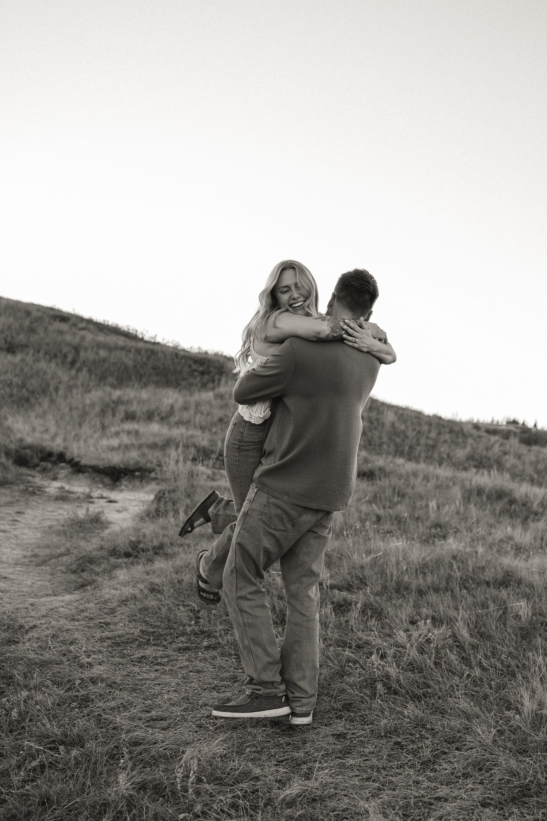 Black-and-white photo of a woman leaping into her partner's arms on a grassy hill.