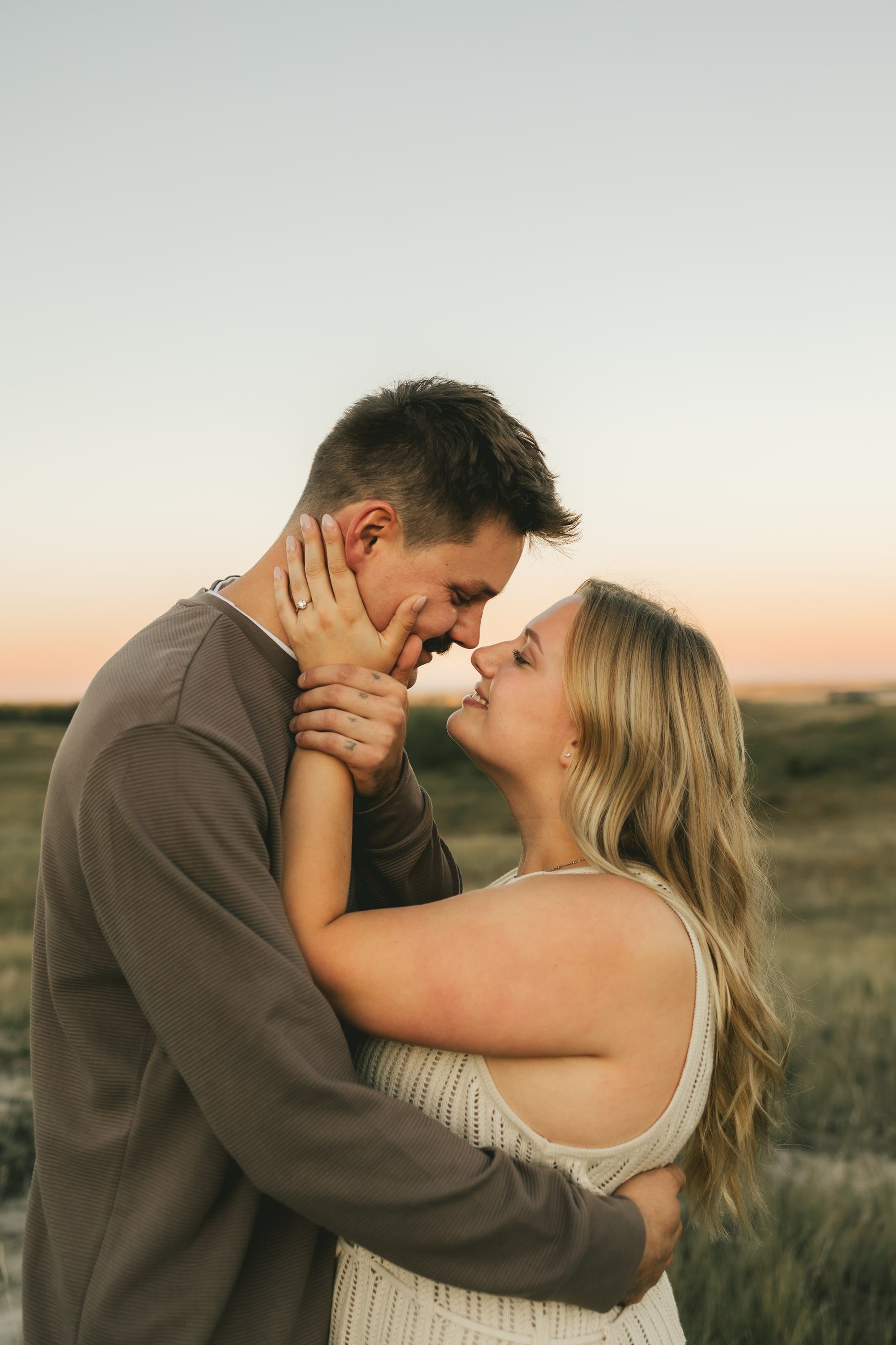 A woman cups her partner's face as they lean in to kiss at sunset.