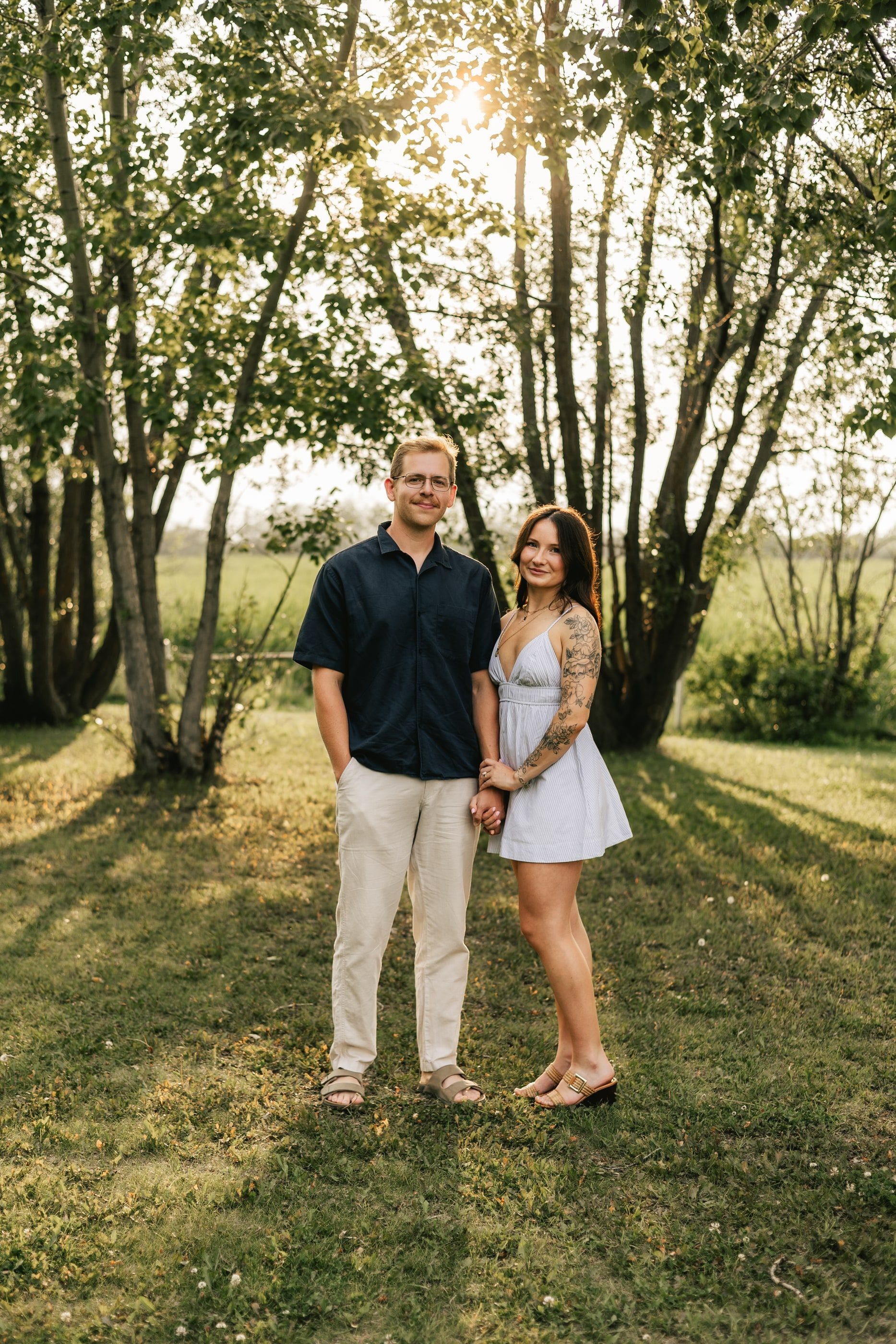 A couple stands side by side under sunlit trees, smiling at the camera.