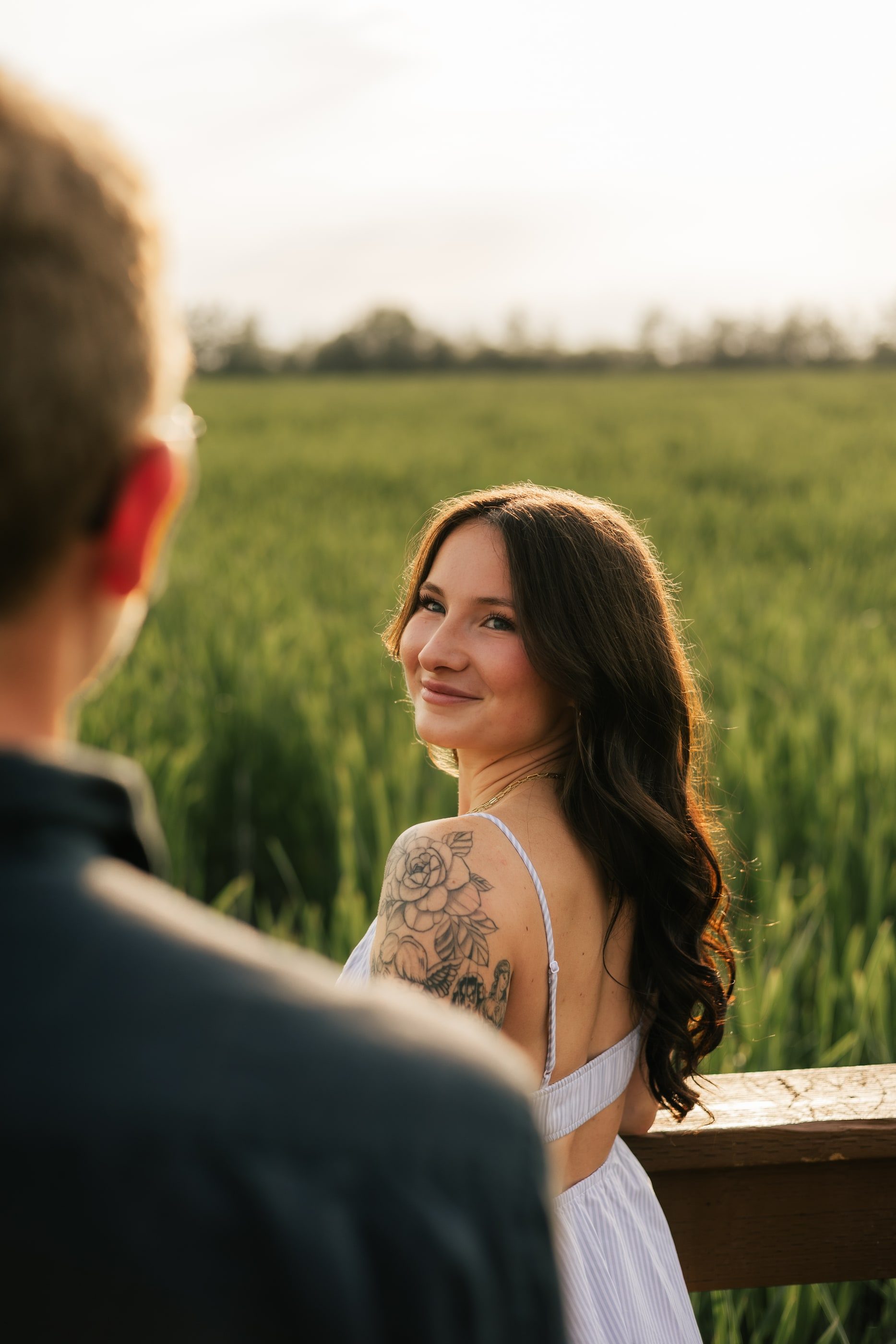 A woman in a white sundress looks back over her shoulder at her partner beside a green field.