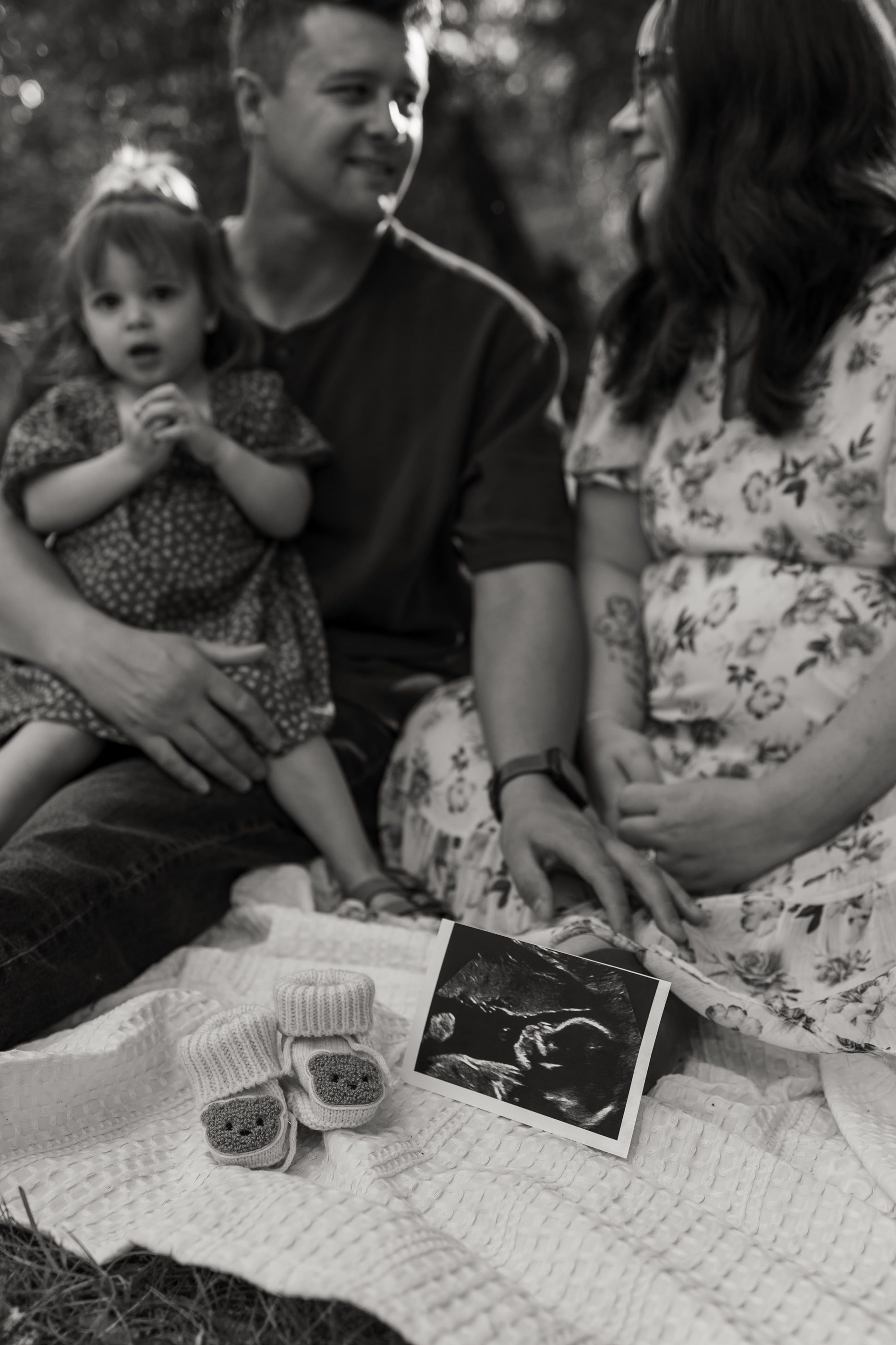 Black-and-white photo of parents and their toddler on a blanket with an ultrasound photo and tiny baby shoes.