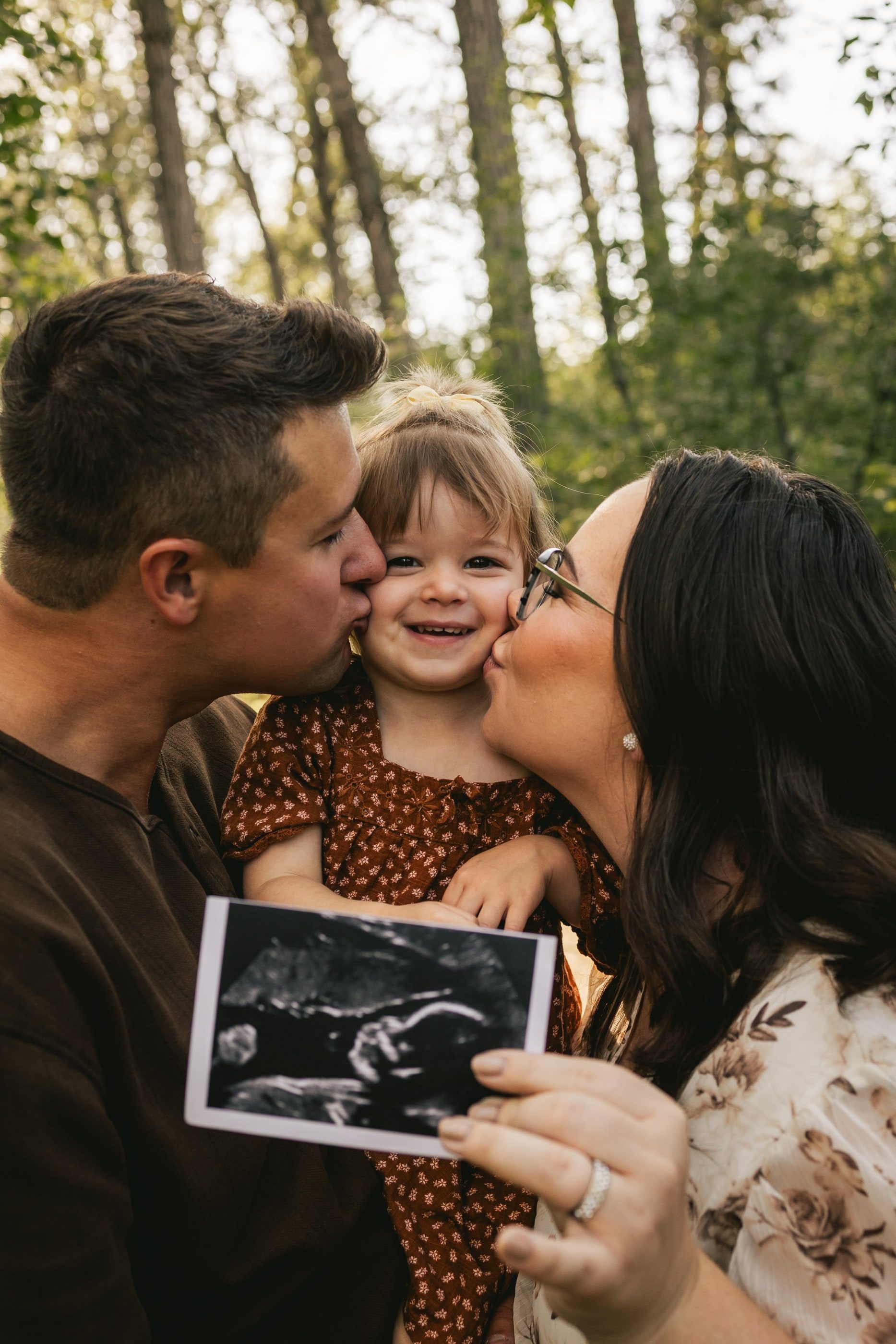 Parents kiss their laughing toddler's cheeks as she holds up an ultrasound photo.
