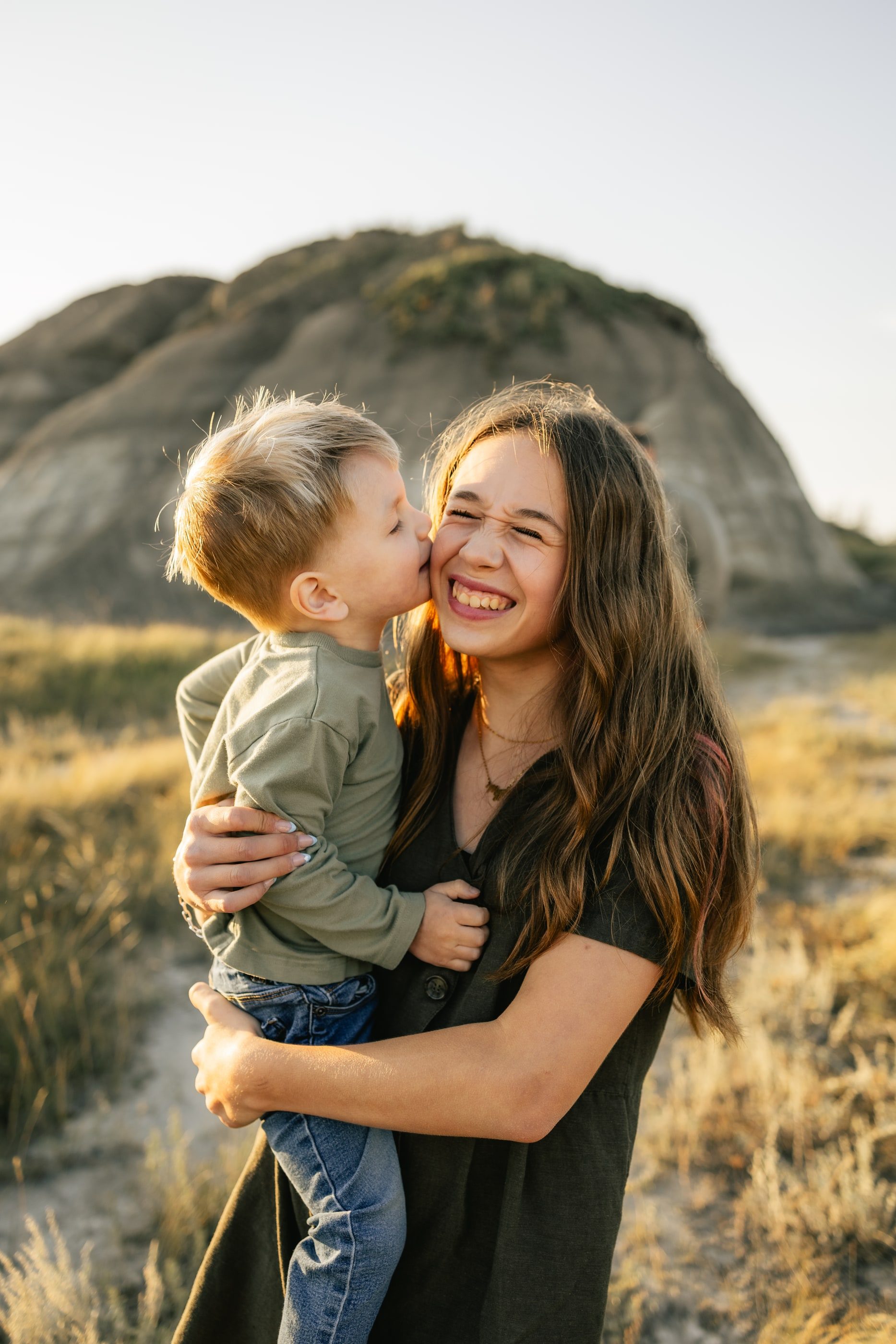 A little boy kisses his laughing mother's cheek as she holds him, badlands hills behind them.