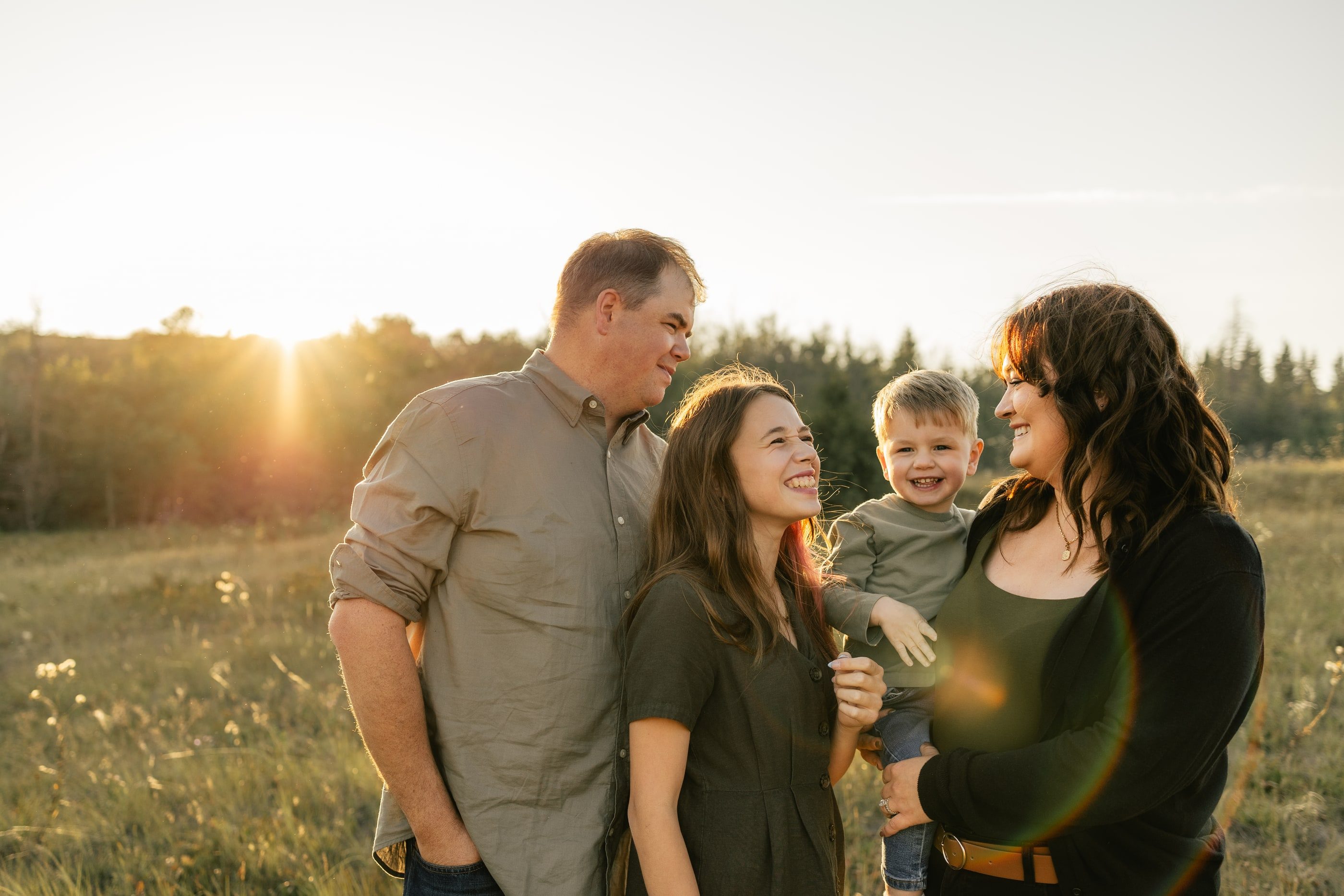 A family of four laughs together in a field, the setting sun behind them.