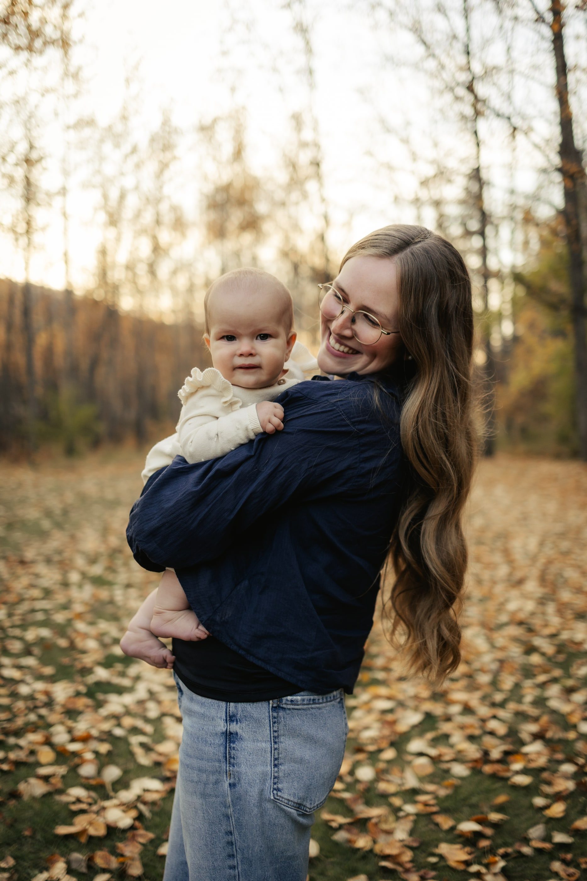A mother laughs as she holds her baby on her hip among fallen autumn leaves.