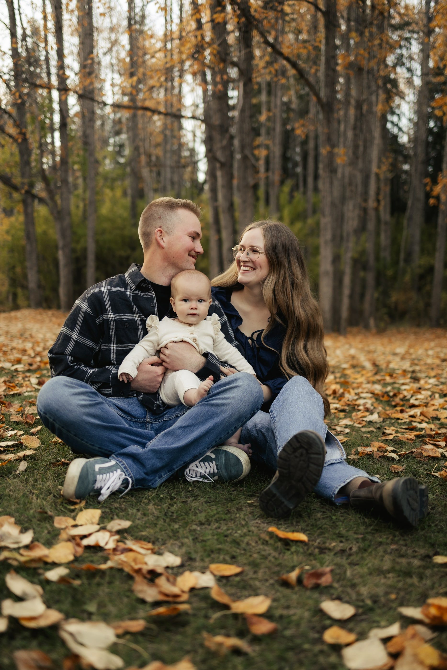 Parents sit with their baby on a carpet of autumn leaves in the forest.