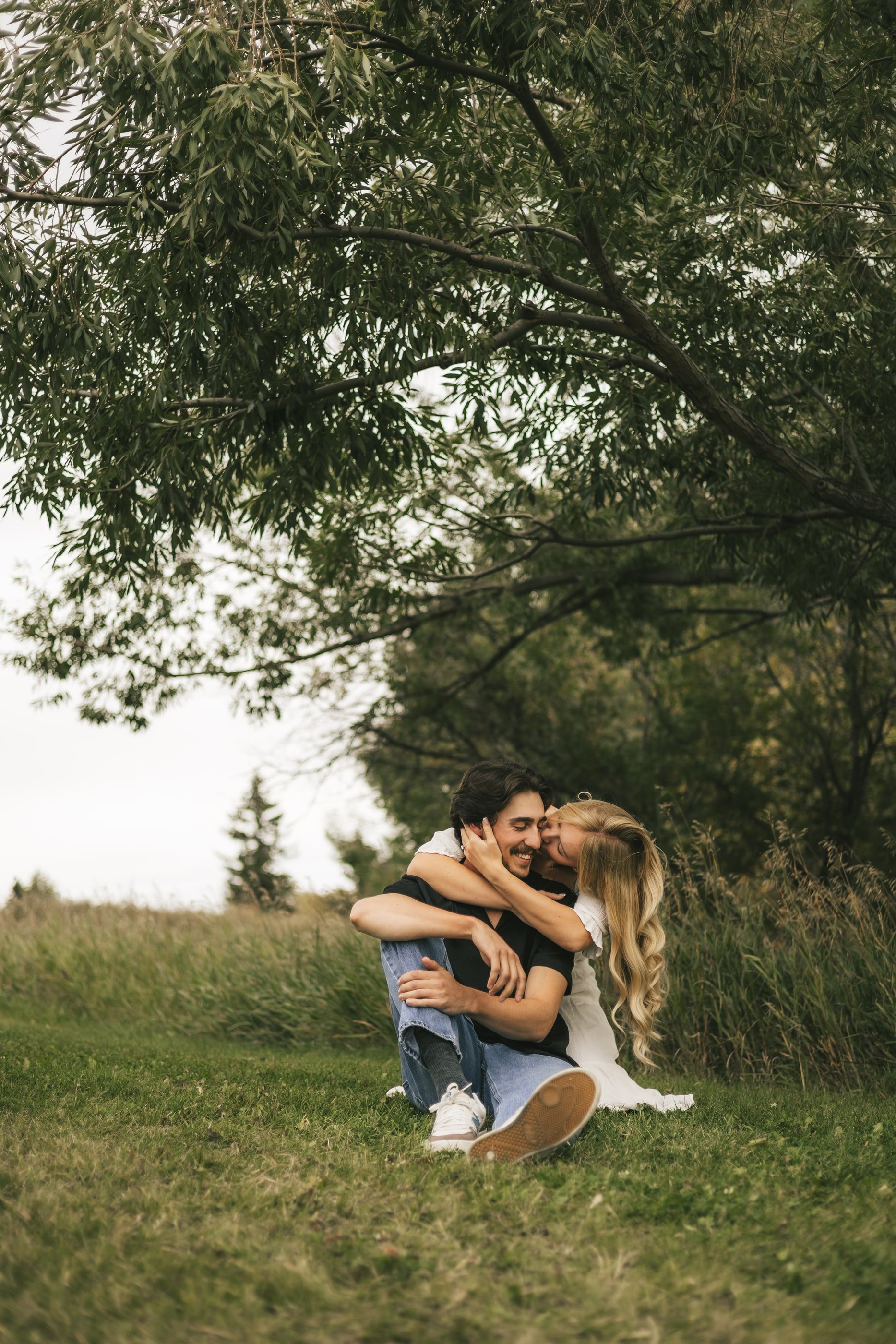 A woman wraps her arms around her partner as they sit laughing in the grass beneath a tree.
