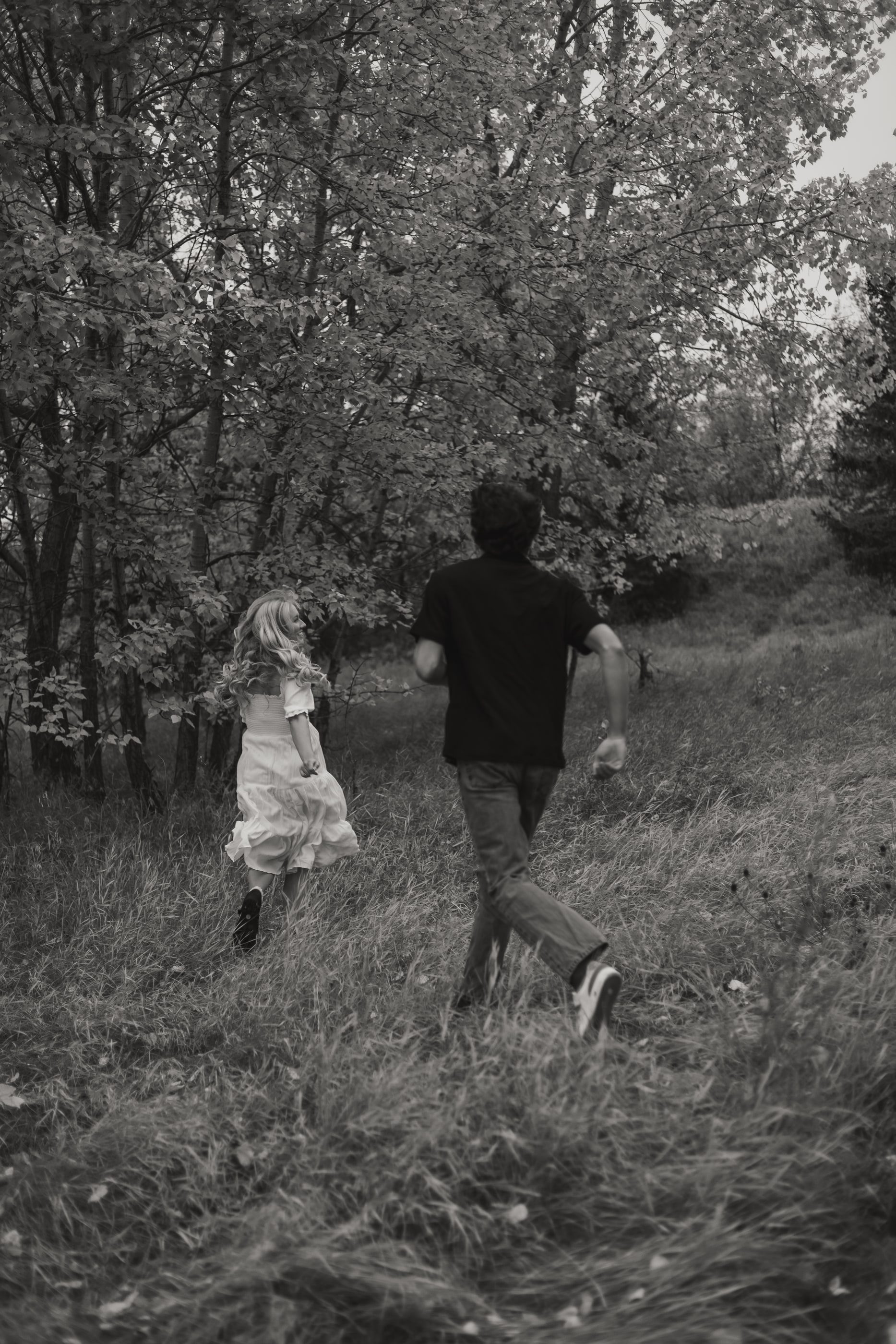 Black-and-white photo of a couple running playfully through long grass toward the trees.