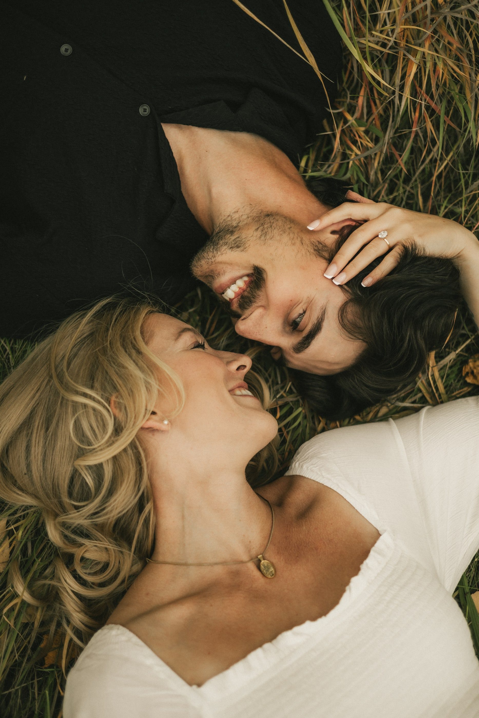 A couple lies head to head in the grass, laughing, her ring visible on her hand.