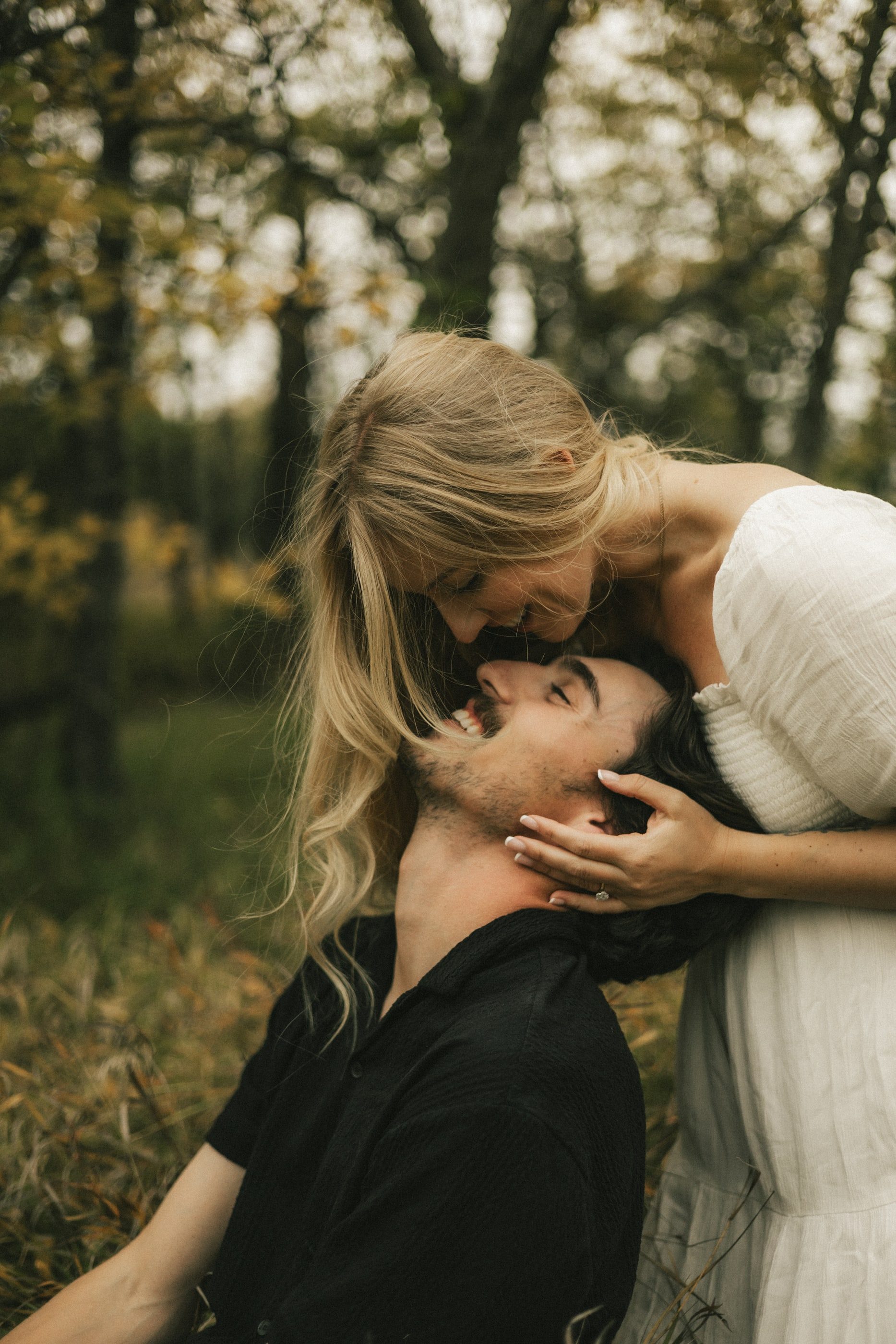 A woman leans down to kiss her partner's forehead as he tilts his head back among the trees.