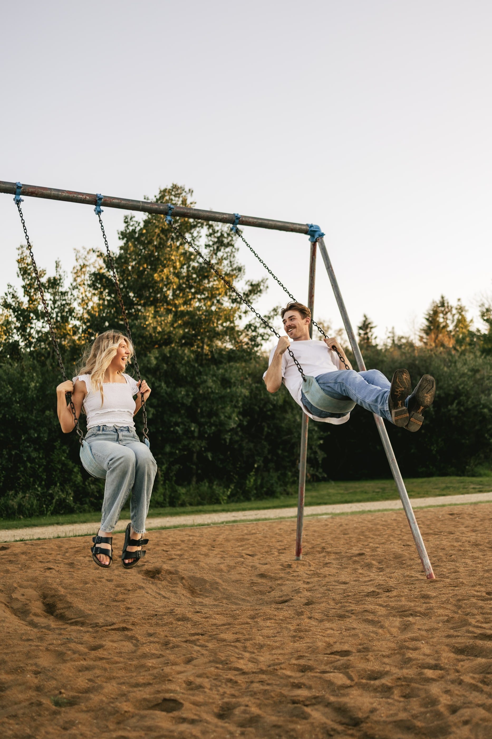 A couple laughs as they swing side by side on a playground swing set at dusk.