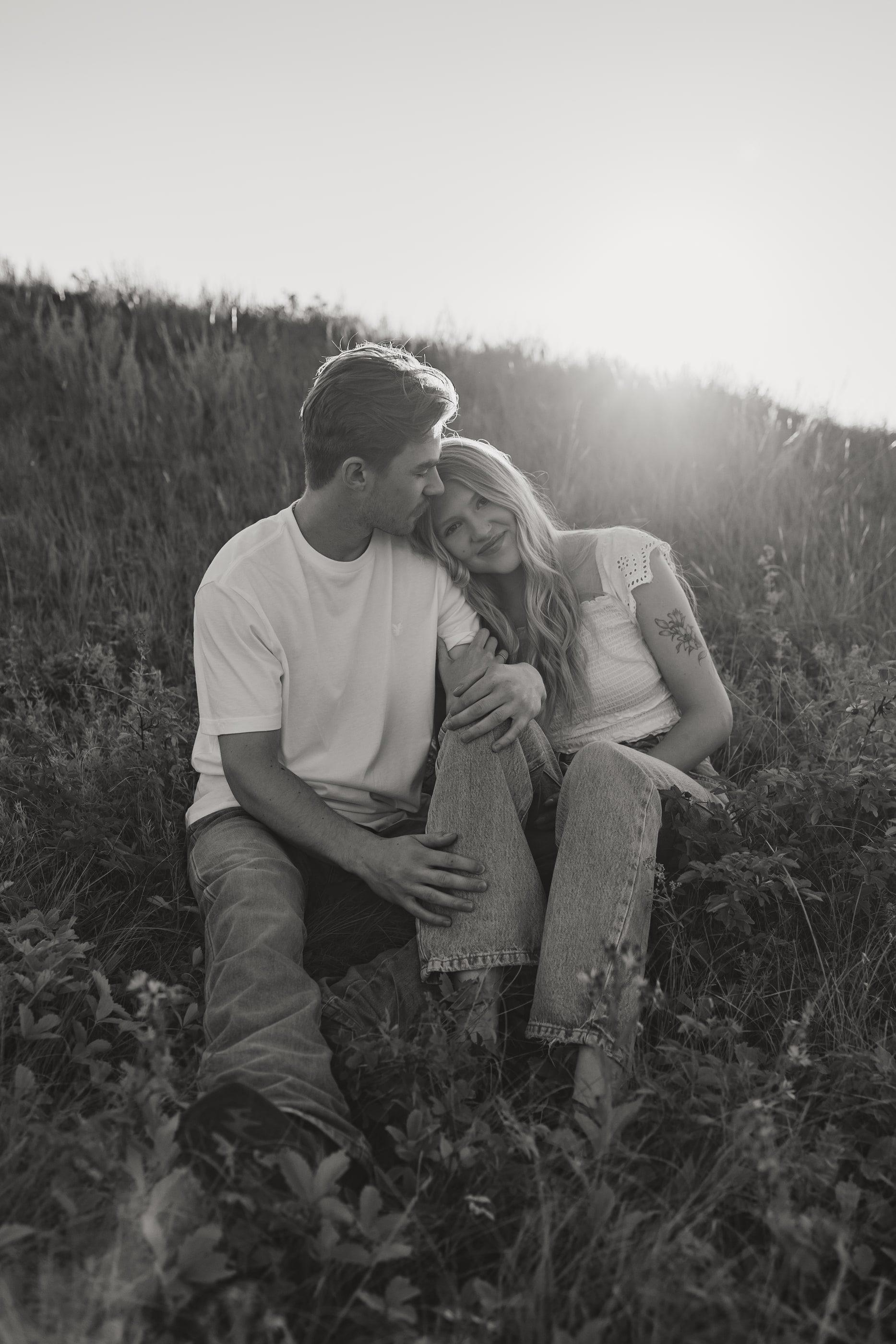 Black-and-white photo of a couple sitting close in tall grass, the sun behind them.