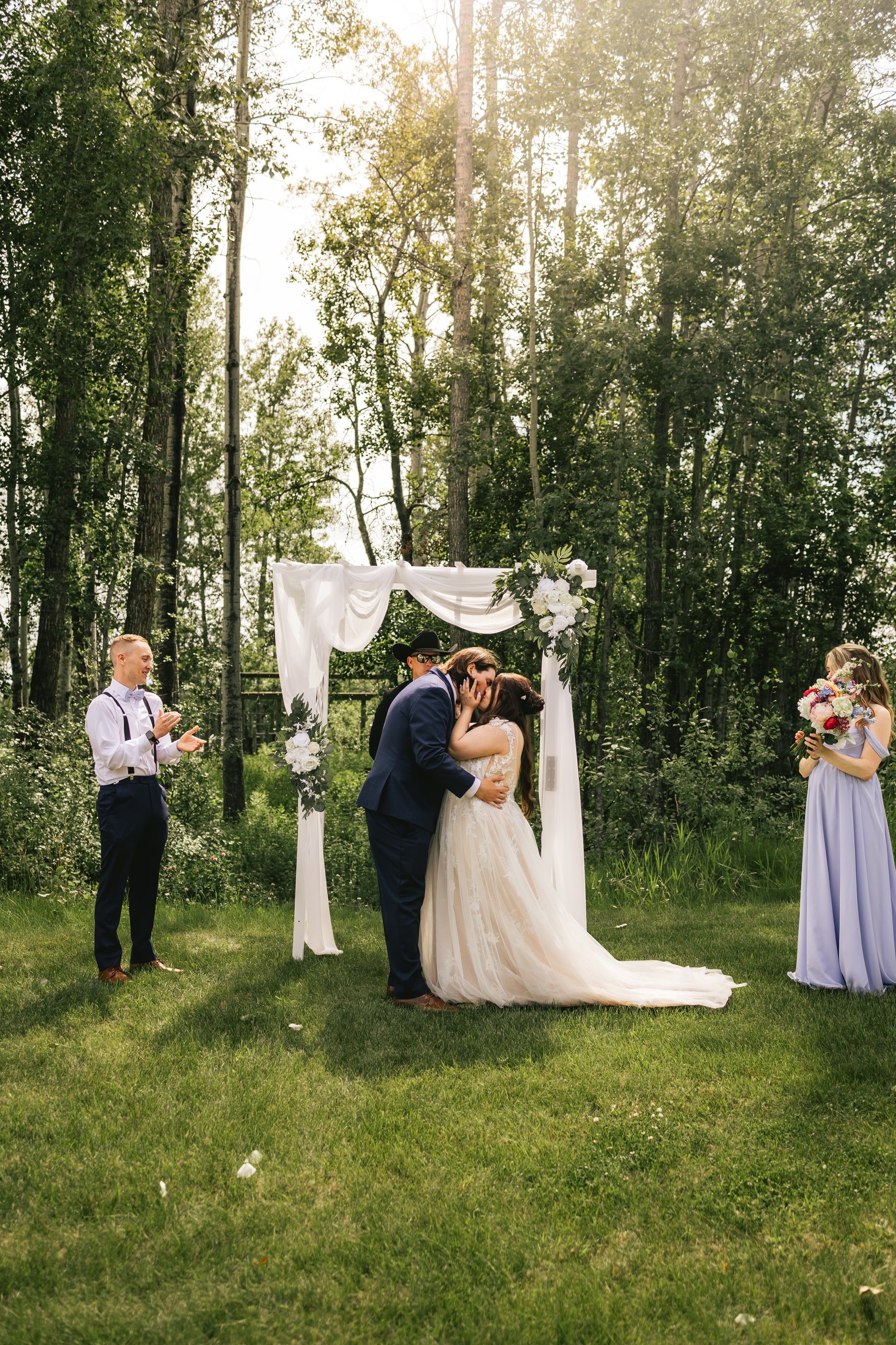 A bride and groom share their first kiss under a white draped arch in the forest.