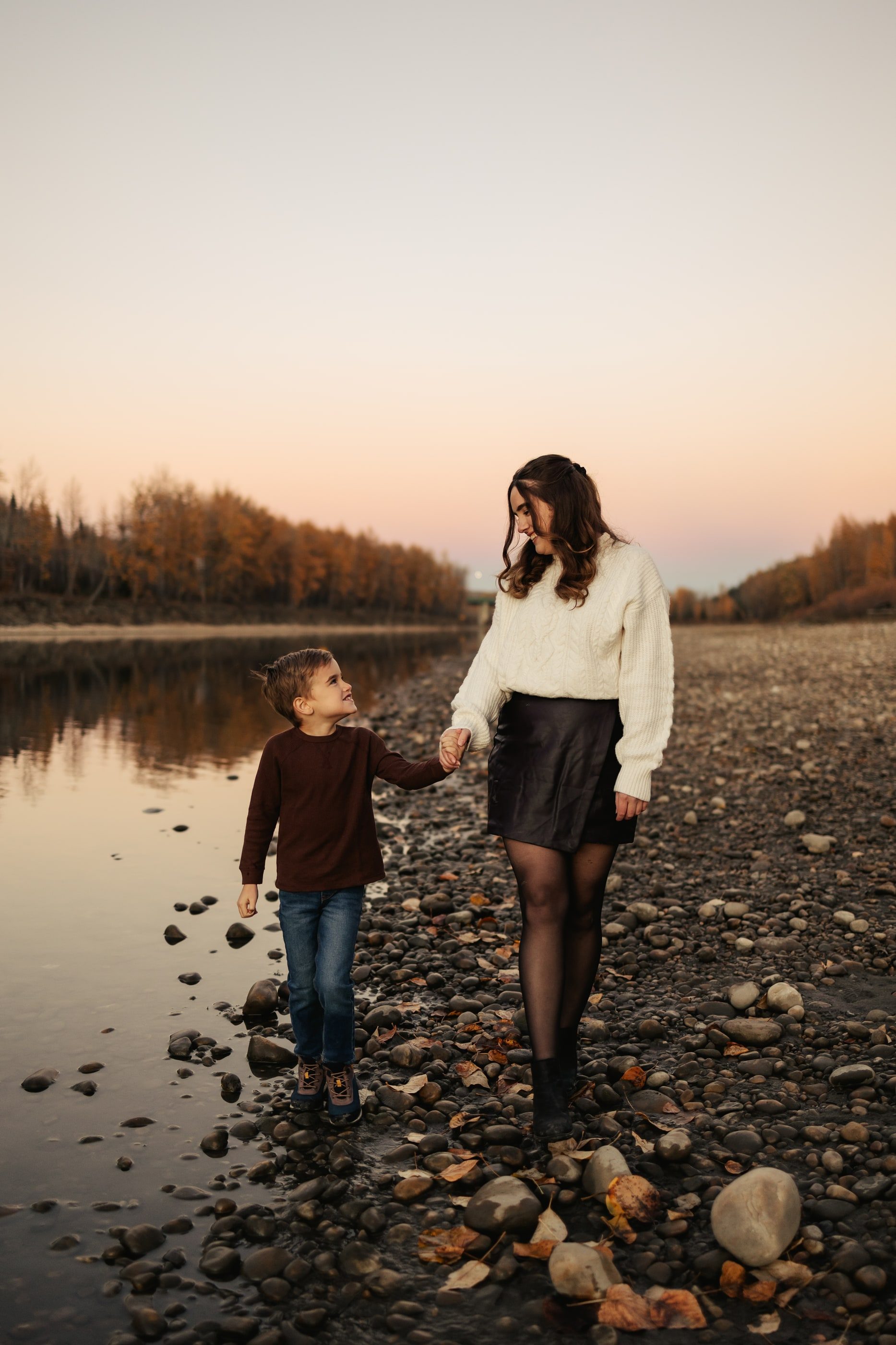 A mother walks hand in hand with her young son along a rocky riverbank at dusk.