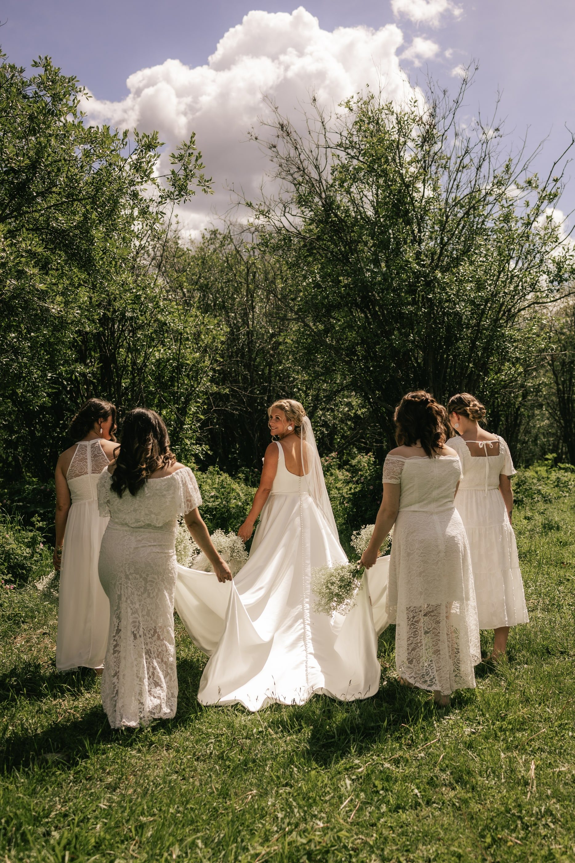 A bride and her bridesmaids in white dresses walk away through a green meadow under summer clouds.