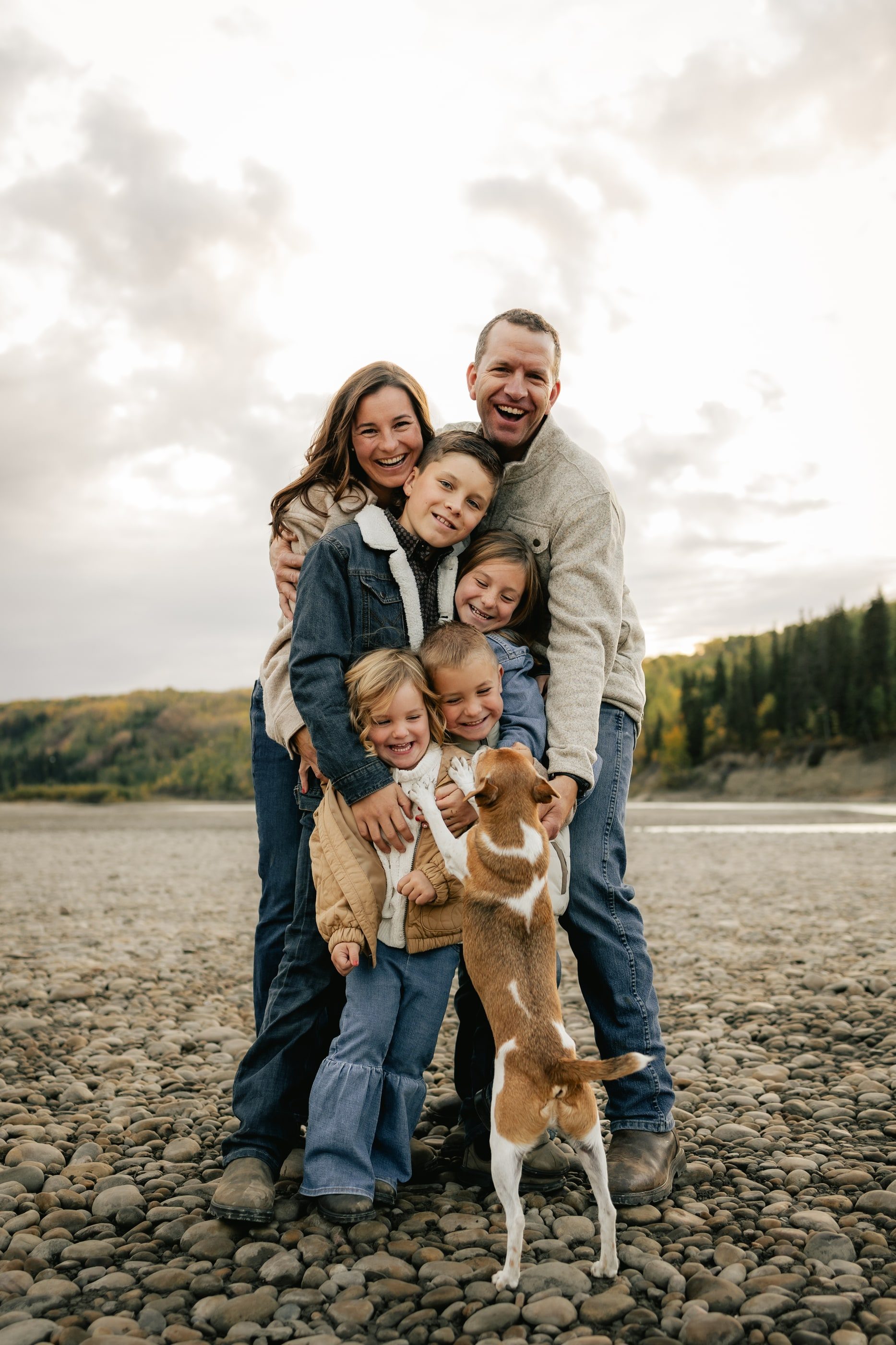 A family of five hugs on a rocky riverbank as their dog jumps up to join in.